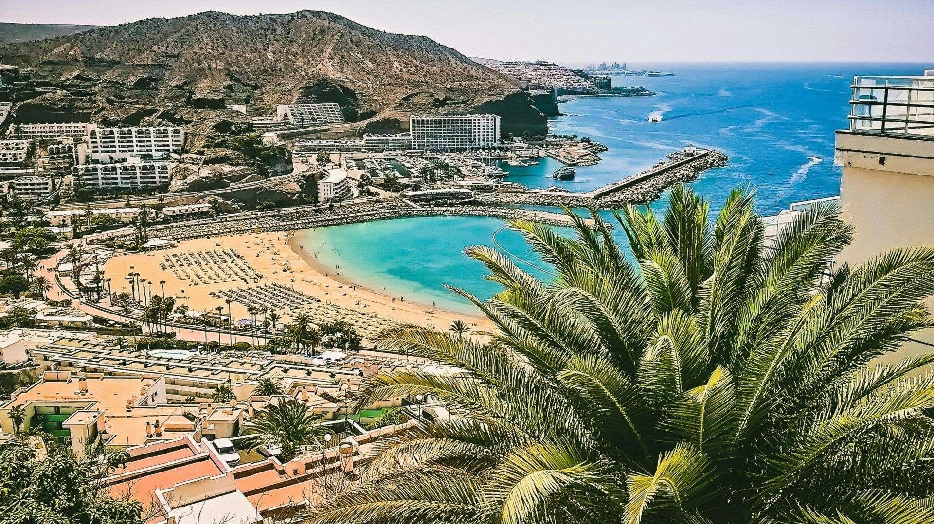 Vista panorámica de una playa con hoteles, montañas rocosas, agua turquesa y palmeras tropicales