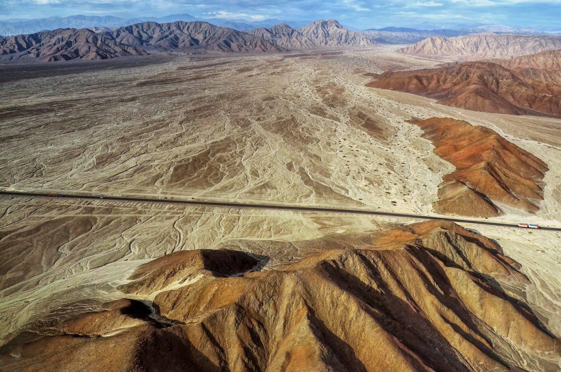 Vista aérea de un valle desértico andino con cadena montañosa, carretera rectilínea y formaciones geológicas de colores
