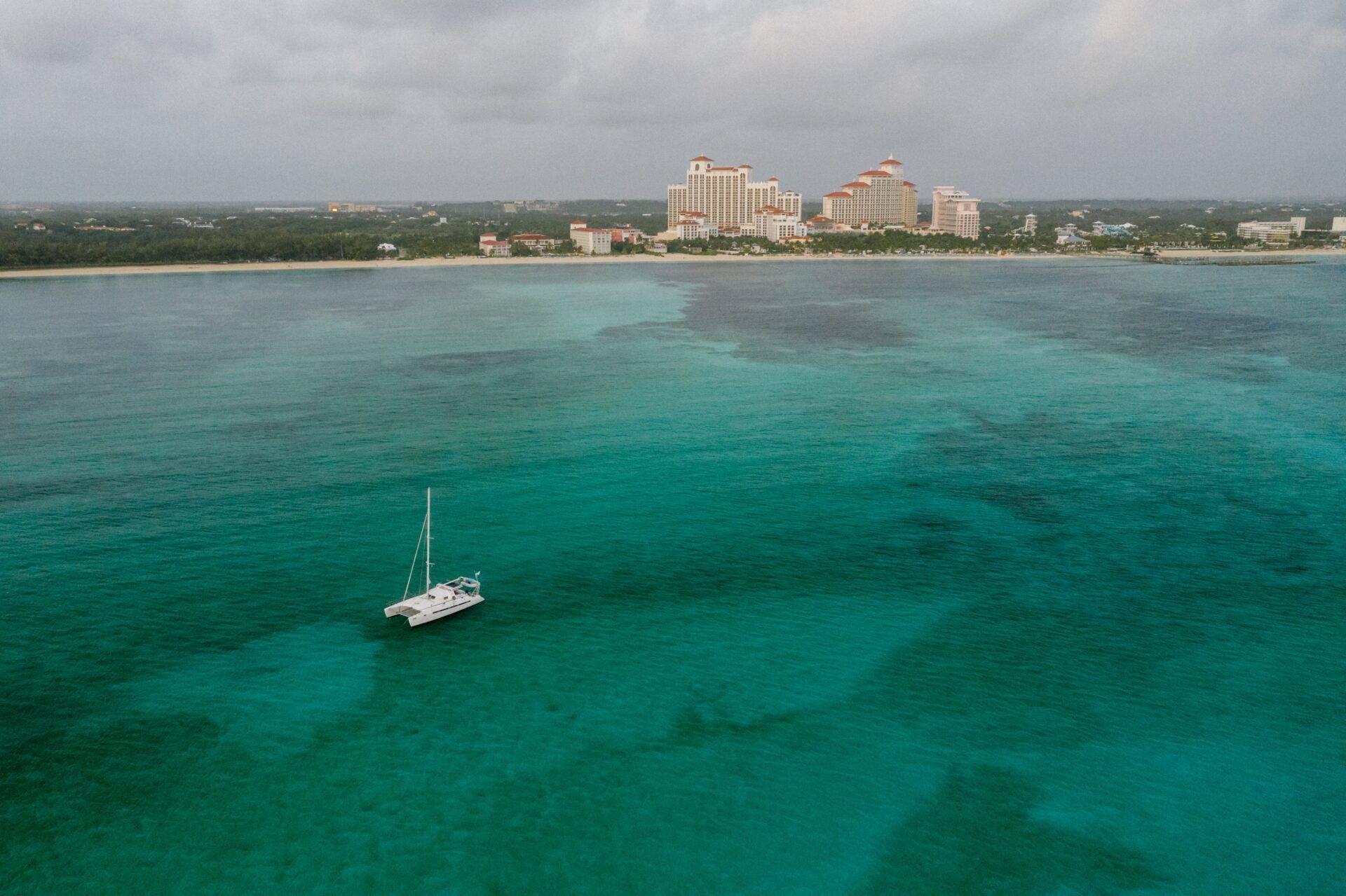 Un velero blanco fondeado en aguas turquesas cristalinas frente a hoteles resort en una costa tropical