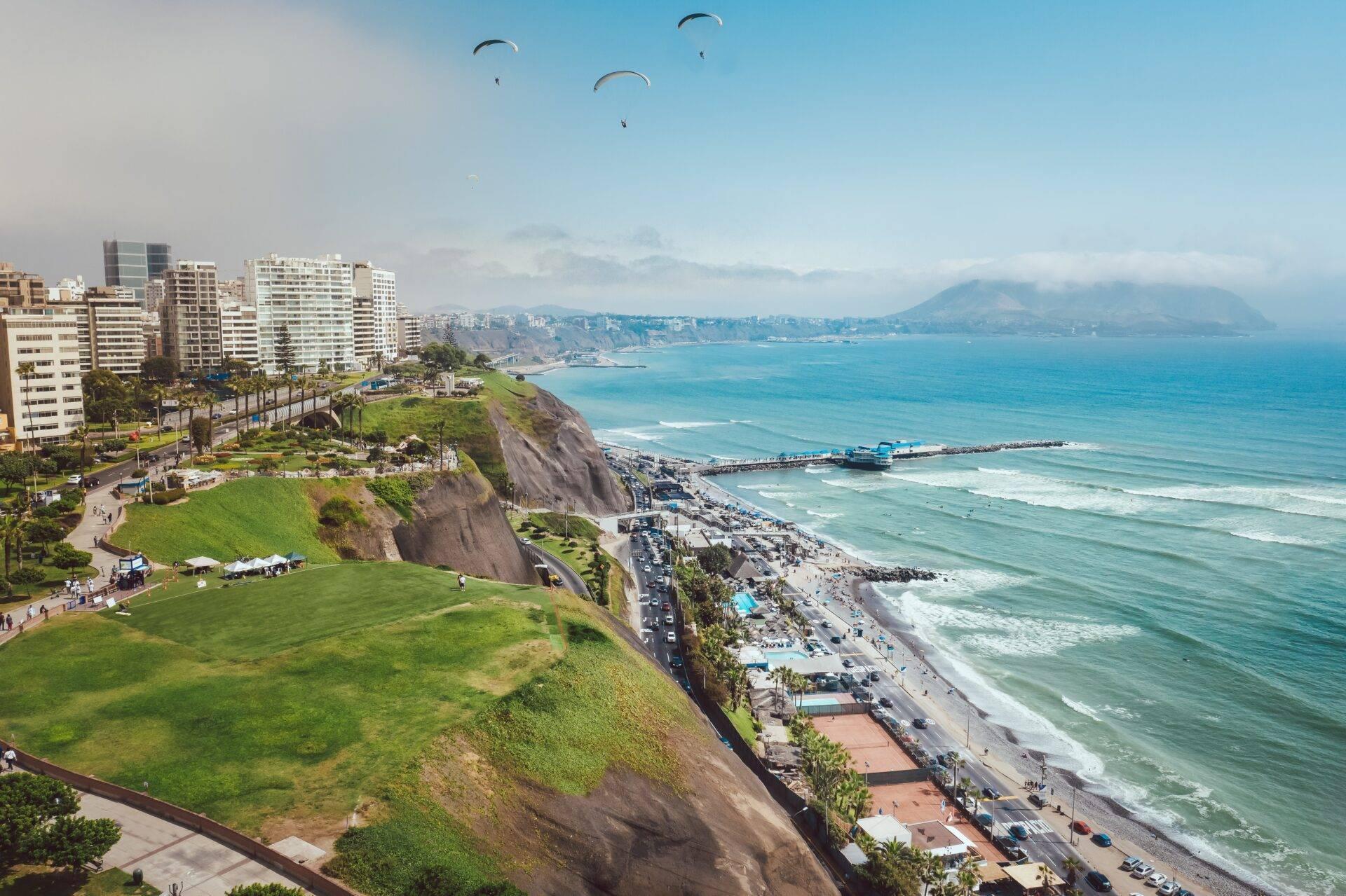 Vista aérea de una costa peruana con acantilados, playa, edificios costeros y parapentistas sobrevolando el océano turquesa