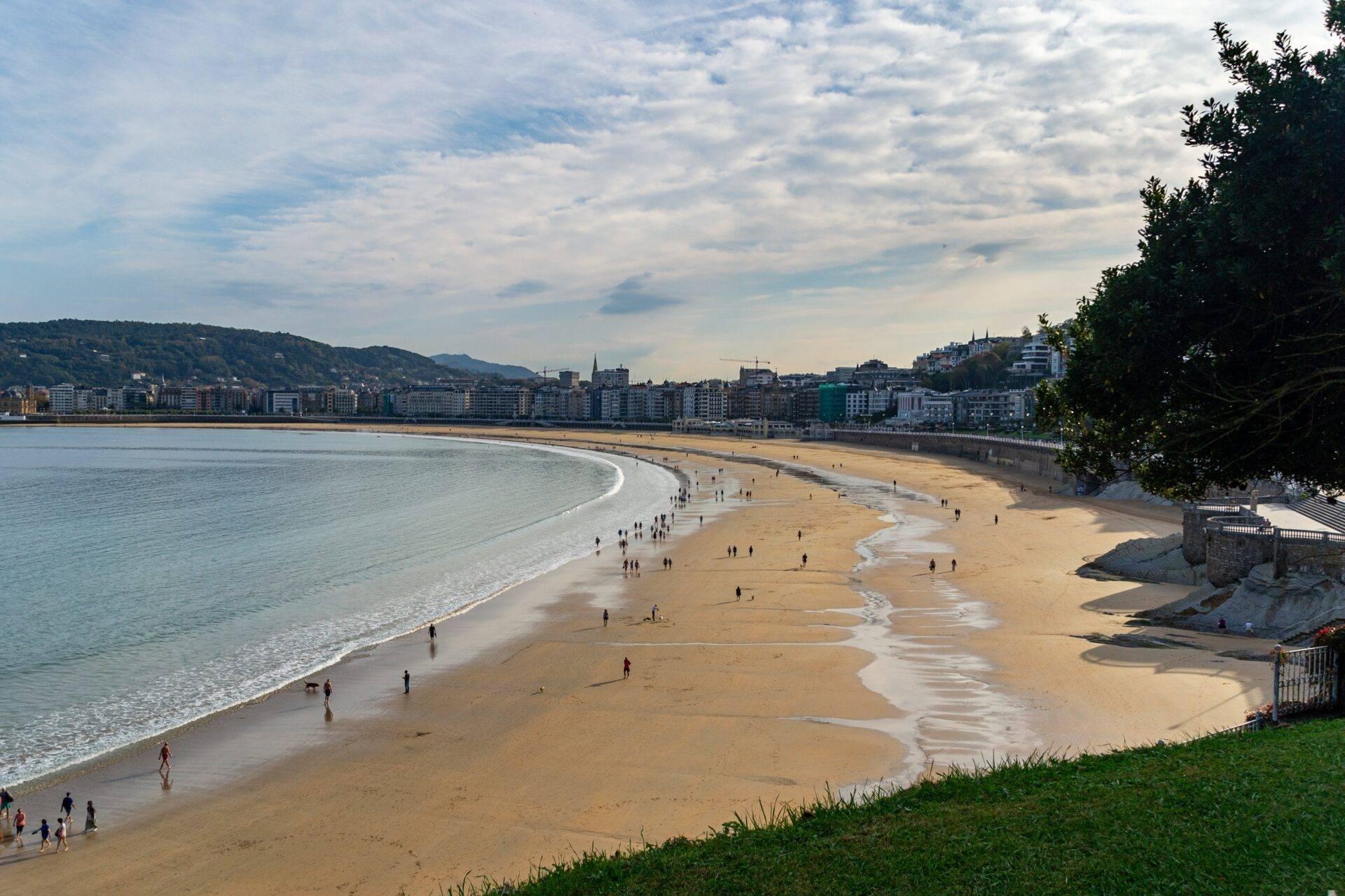 Vista panoramica di una grande spiaggia di sabbia fine con bagnanti e una città costiera sullo sfondo sotto un cielo nuvoloso