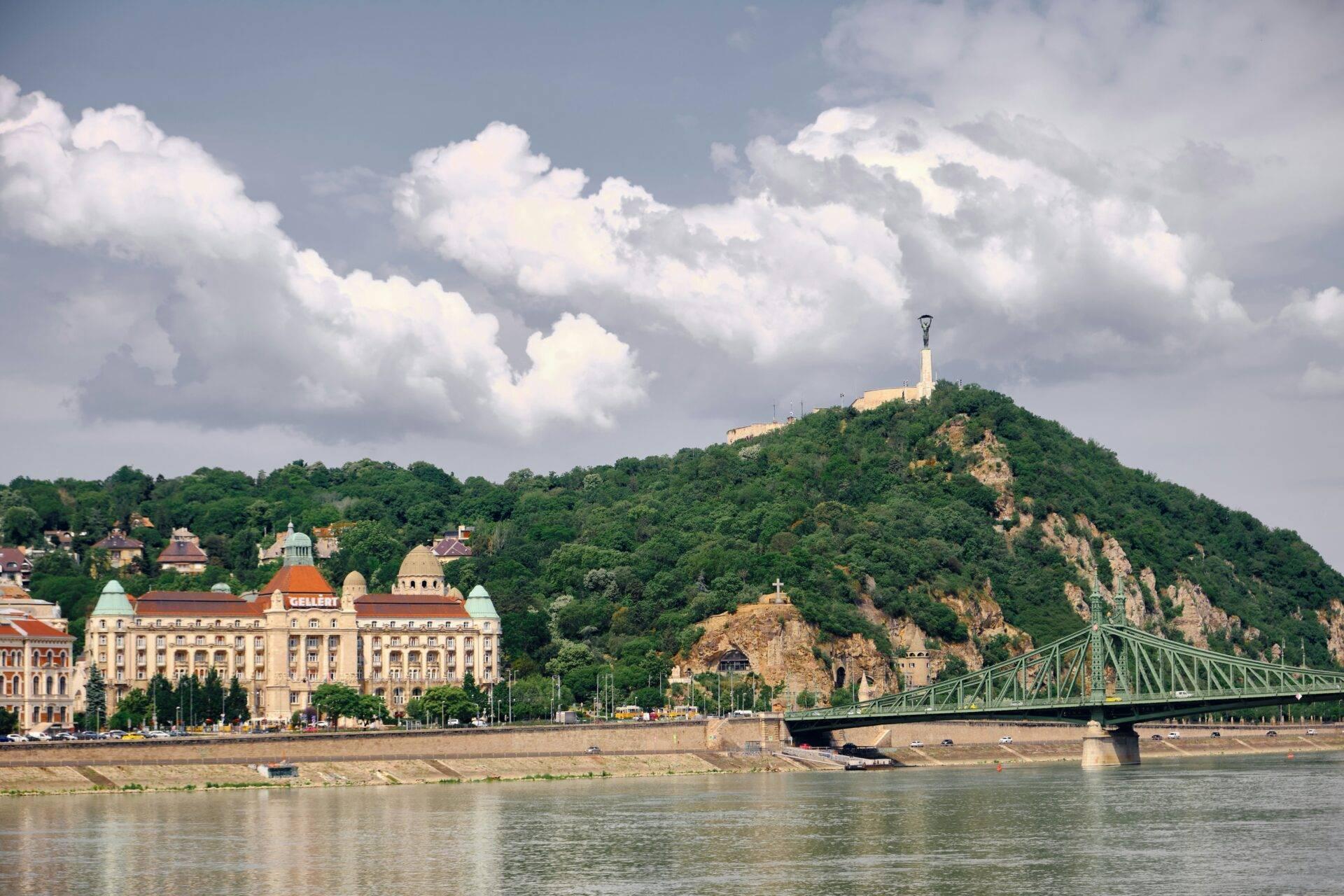 Vista panorámica de una ciudad fluvial con un monumento conmemorativo en lo alto de una colina boscosa y un puente metálico