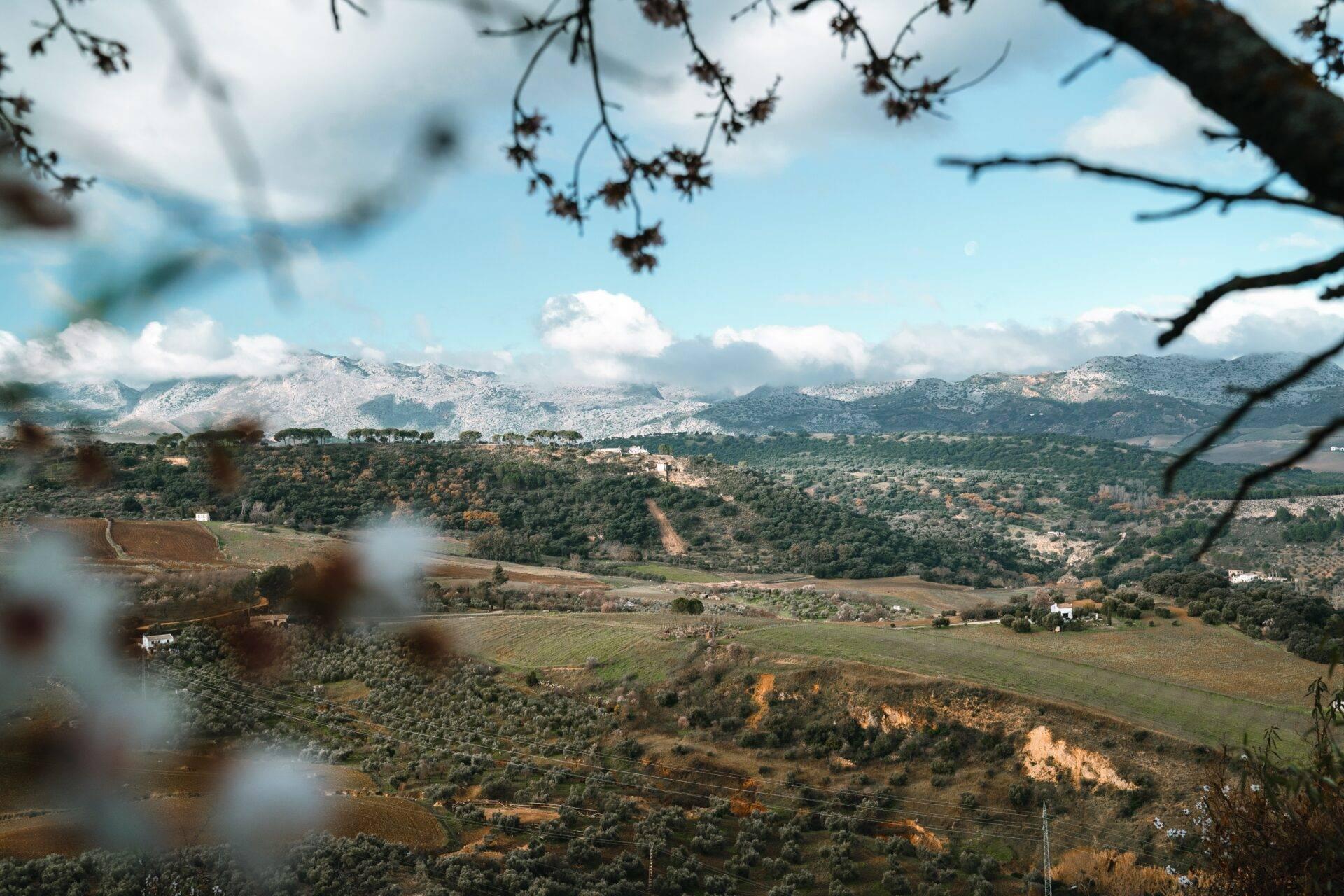 Vista panoramica su una valle mediterranea con montagne innevate, villaggi sparsi e campi coltivati sotto un cielo azzurro
