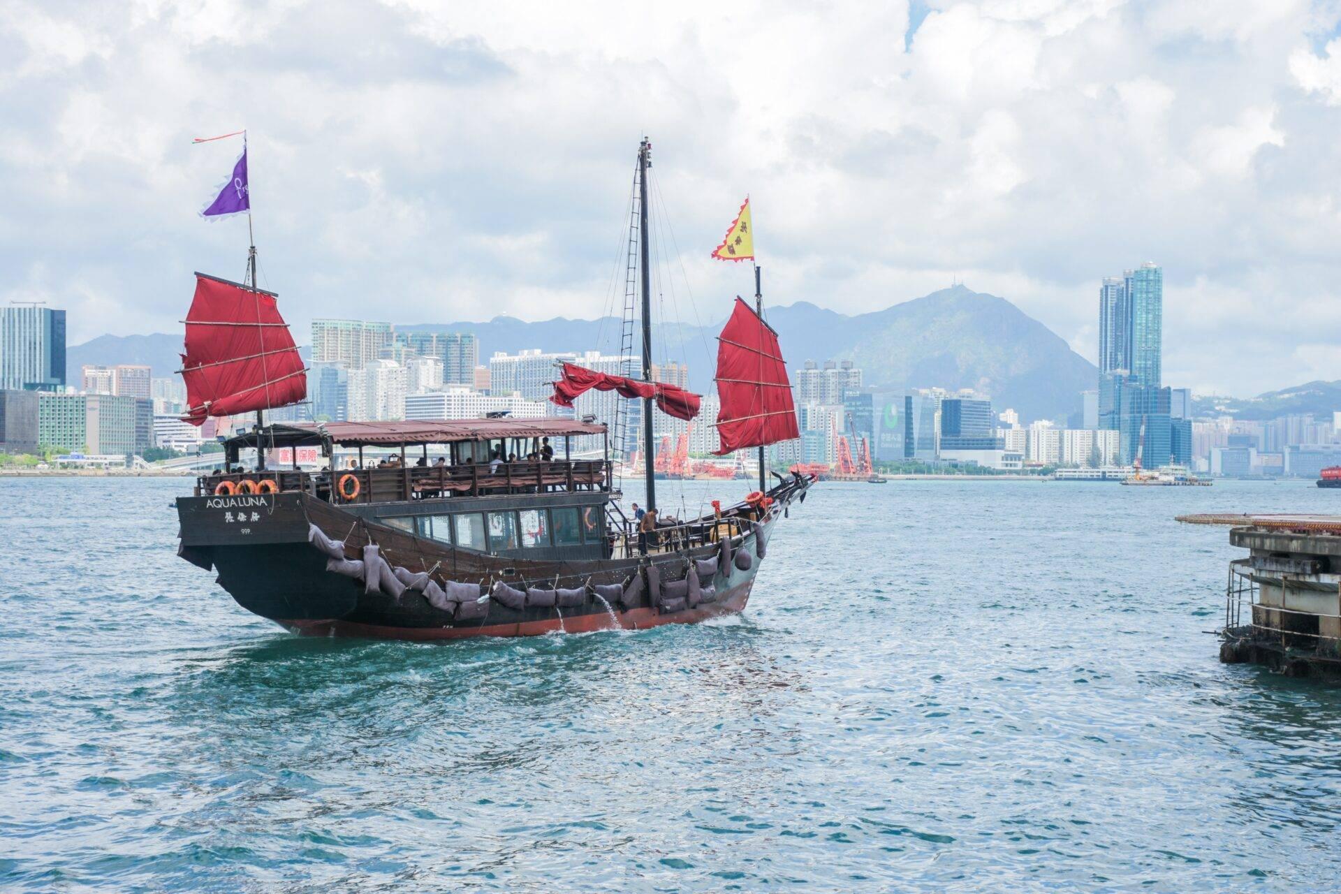 Traditionelle Dschunke mit roten Segeln in einem Stadthafen mit Wolkenkratzern und Bergen im Hintergrund