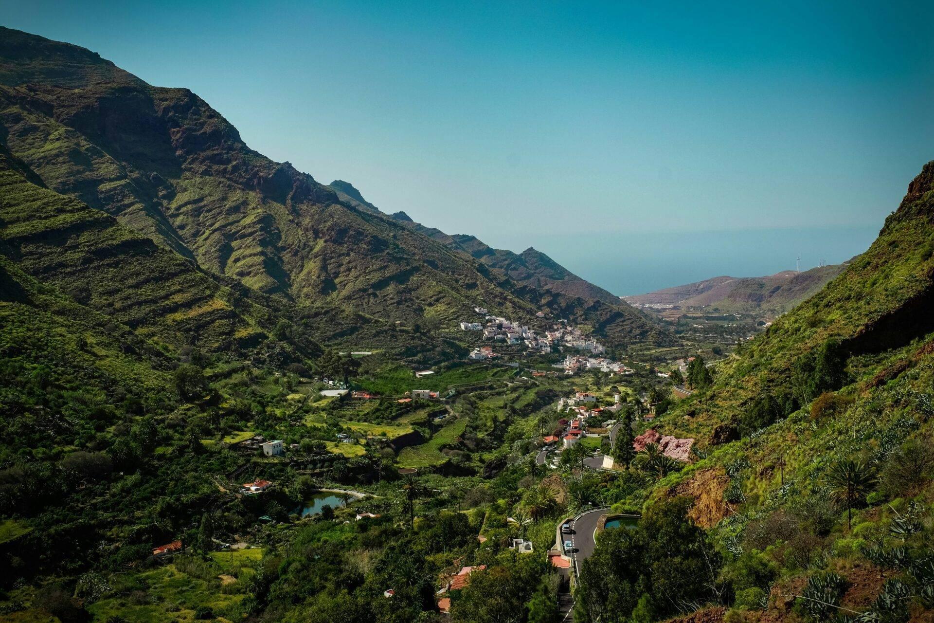 Vista aérea de un valle verde con pueblos blancos, carretera serpenteante y montañas escarpadas bajo cielo despejado