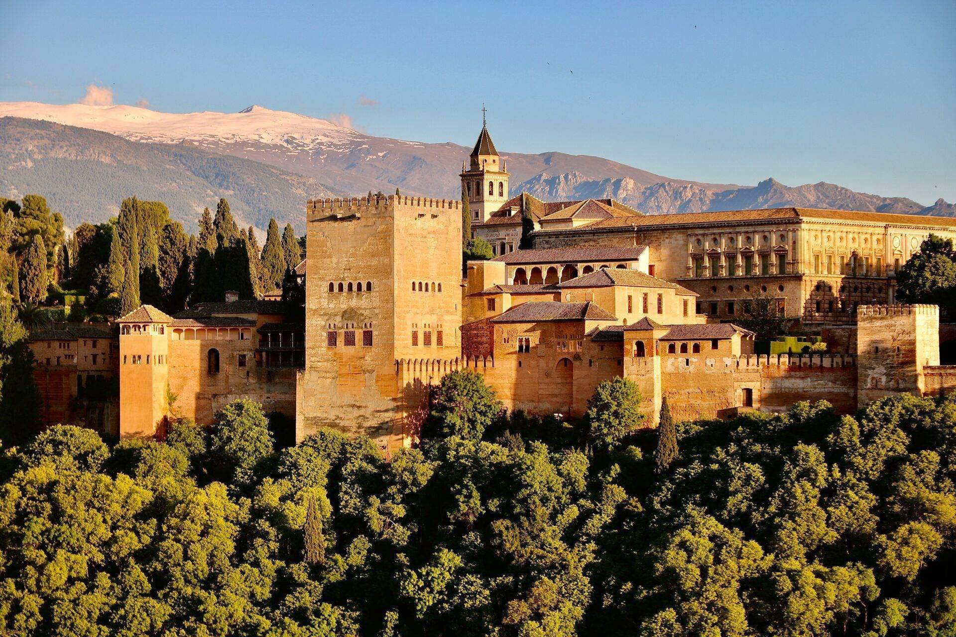 Fortezza medievale dorata con torri e mura, circondata da vegetazione verdeggiante e montagne innevate sullo sfondo