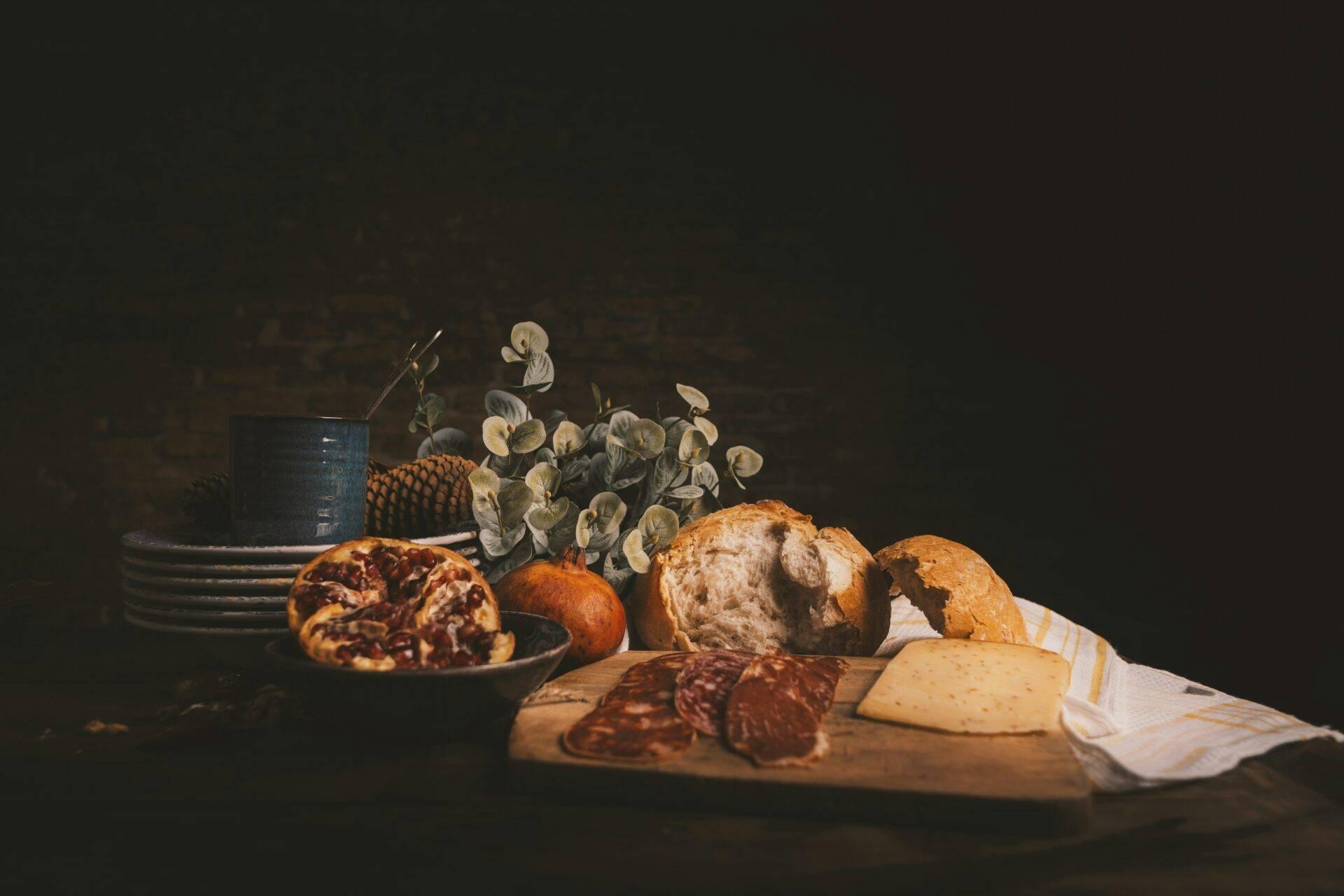 Composición culinaria con pan, embutido, queso, frutas y flores secas sobre fondo oscuro