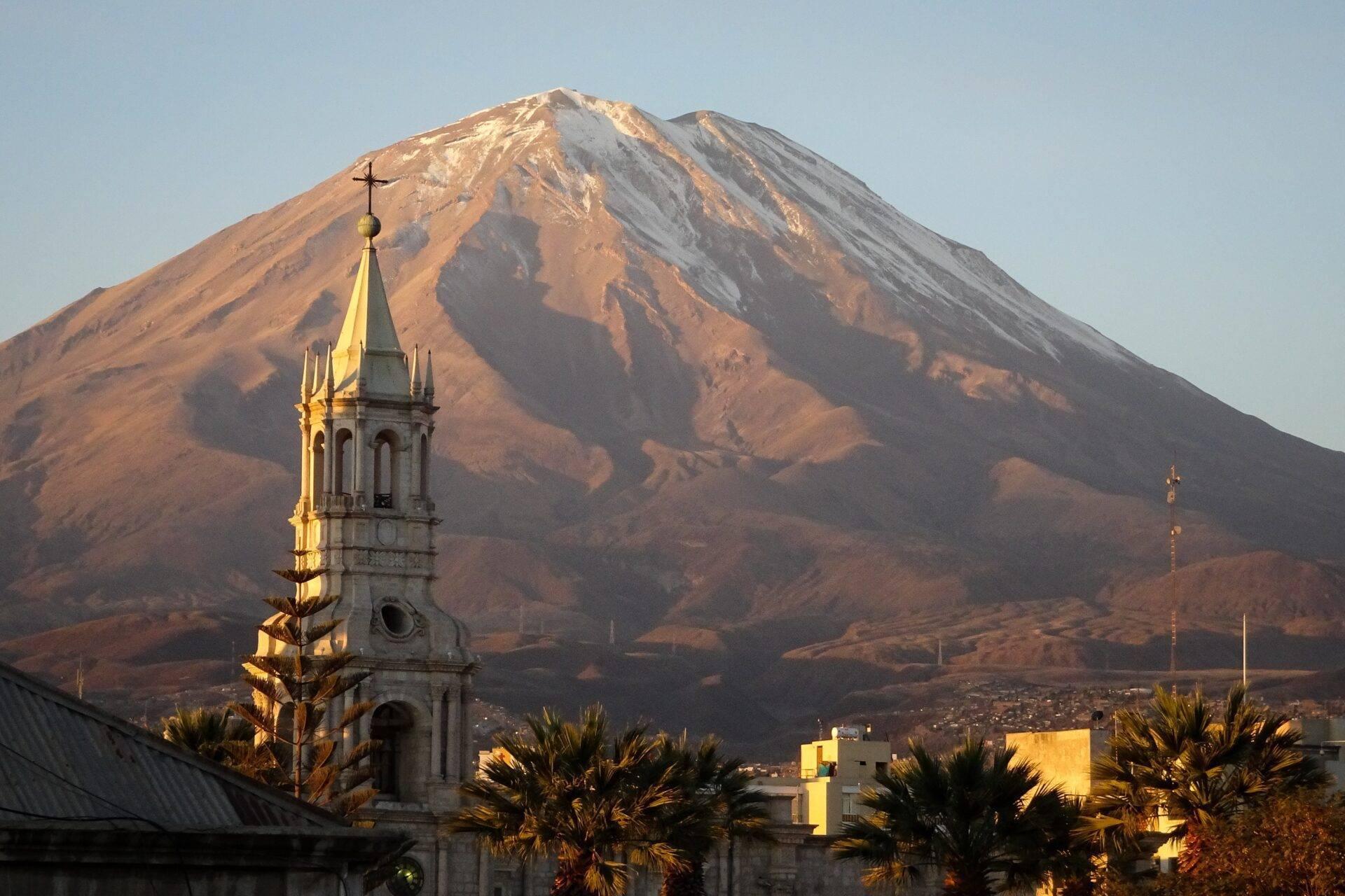 Un campanario blanco con cruz dorada frente a un volcán nevado en los Andes peruanos