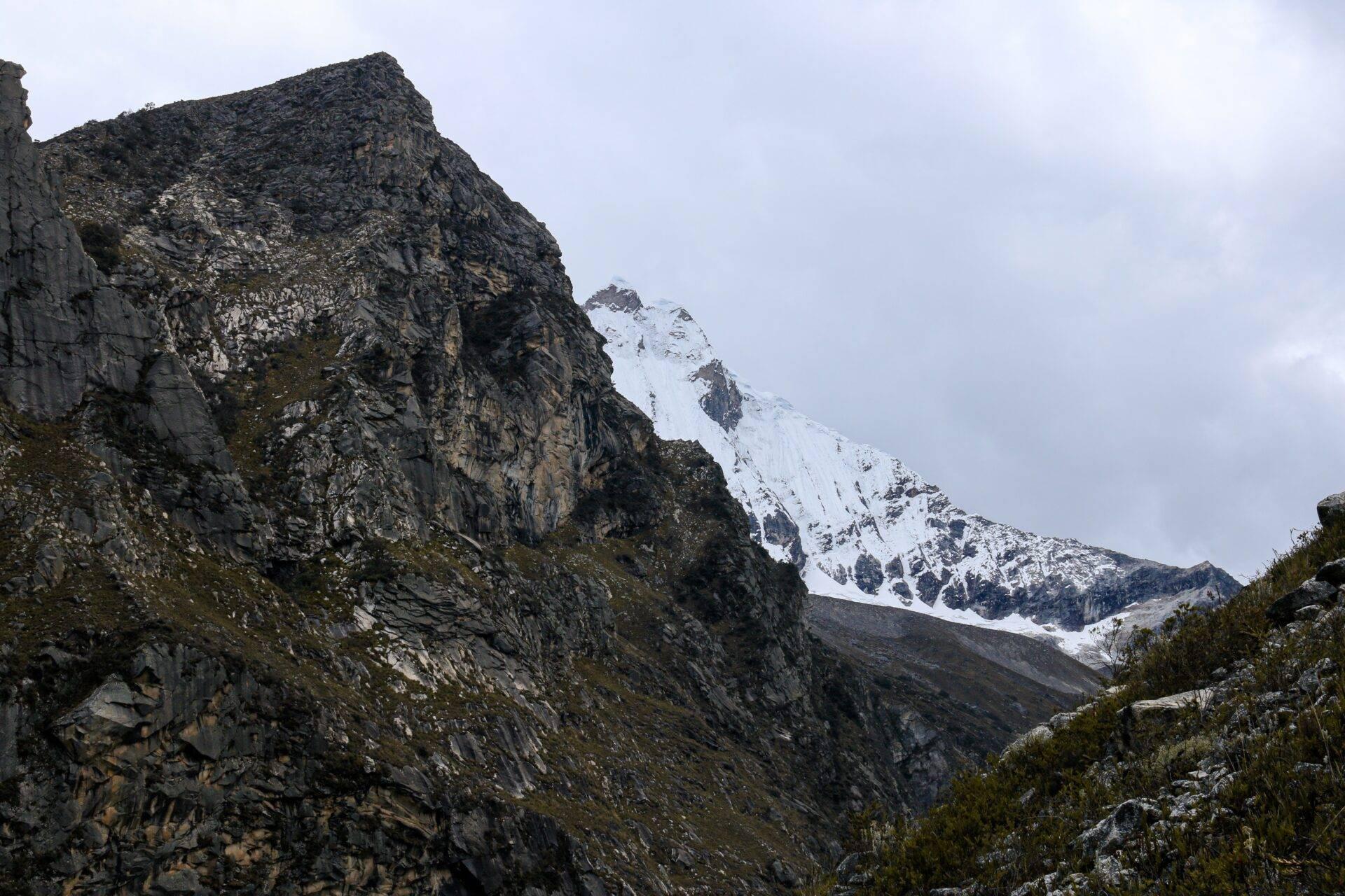 Vista de un valle alpino con picos rocosos escarpados y montaña nevada al fondo bajo cielo nublado