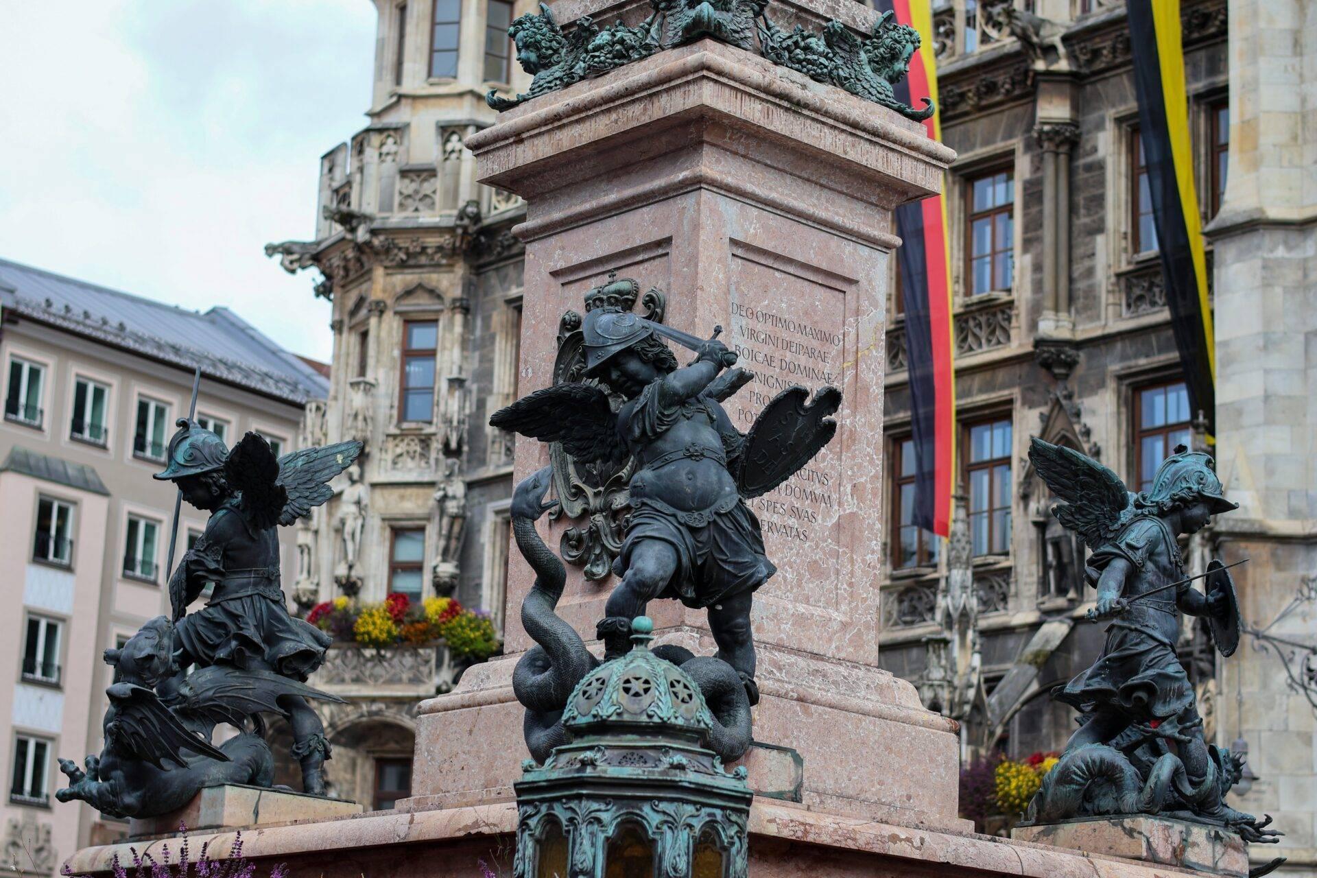Monumento con statue di angeli alati in bronzo su piedistallo di pietra in una piazza storica europea circondata da architettura classica