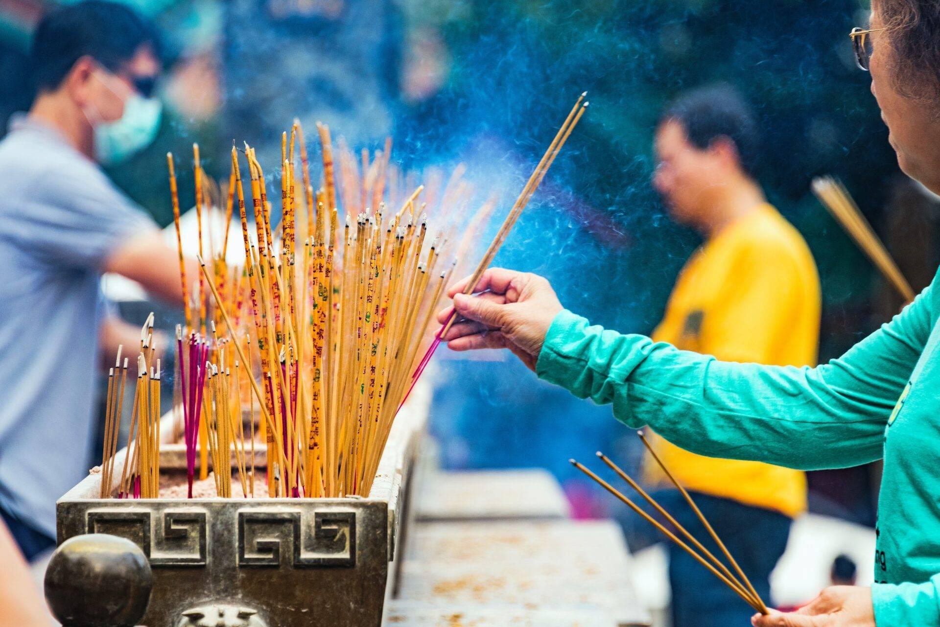 Person in grüner Jacke hält ein Räucherstäbchen an einem verzierten Weihrauchbrenner, umgeben von Besuchern in einem asiatischen Tempel