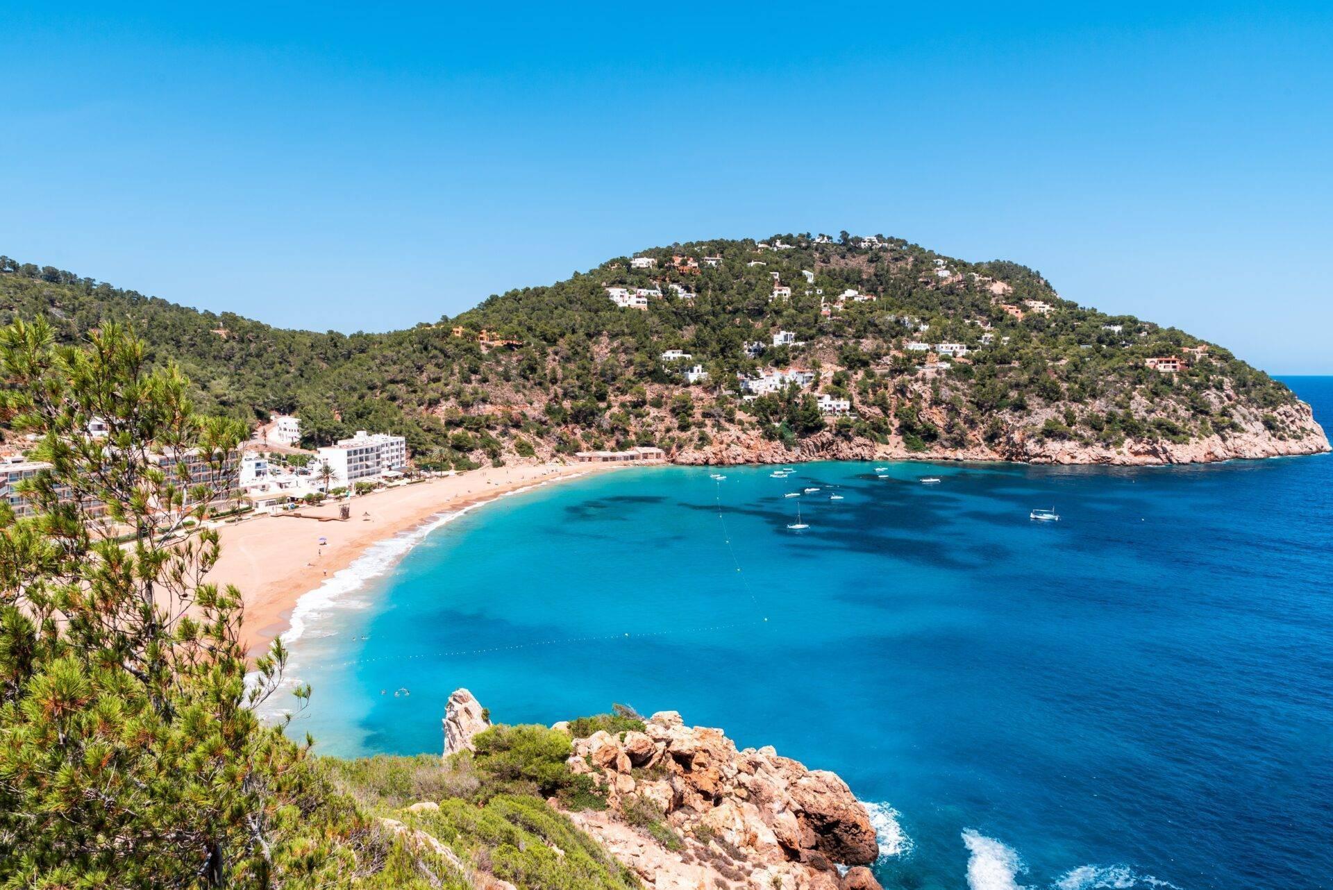 Una baia mediterranea con acque turchesi, spiaggia di sabbia fine e scogliere rocciose boscose con abitazioni