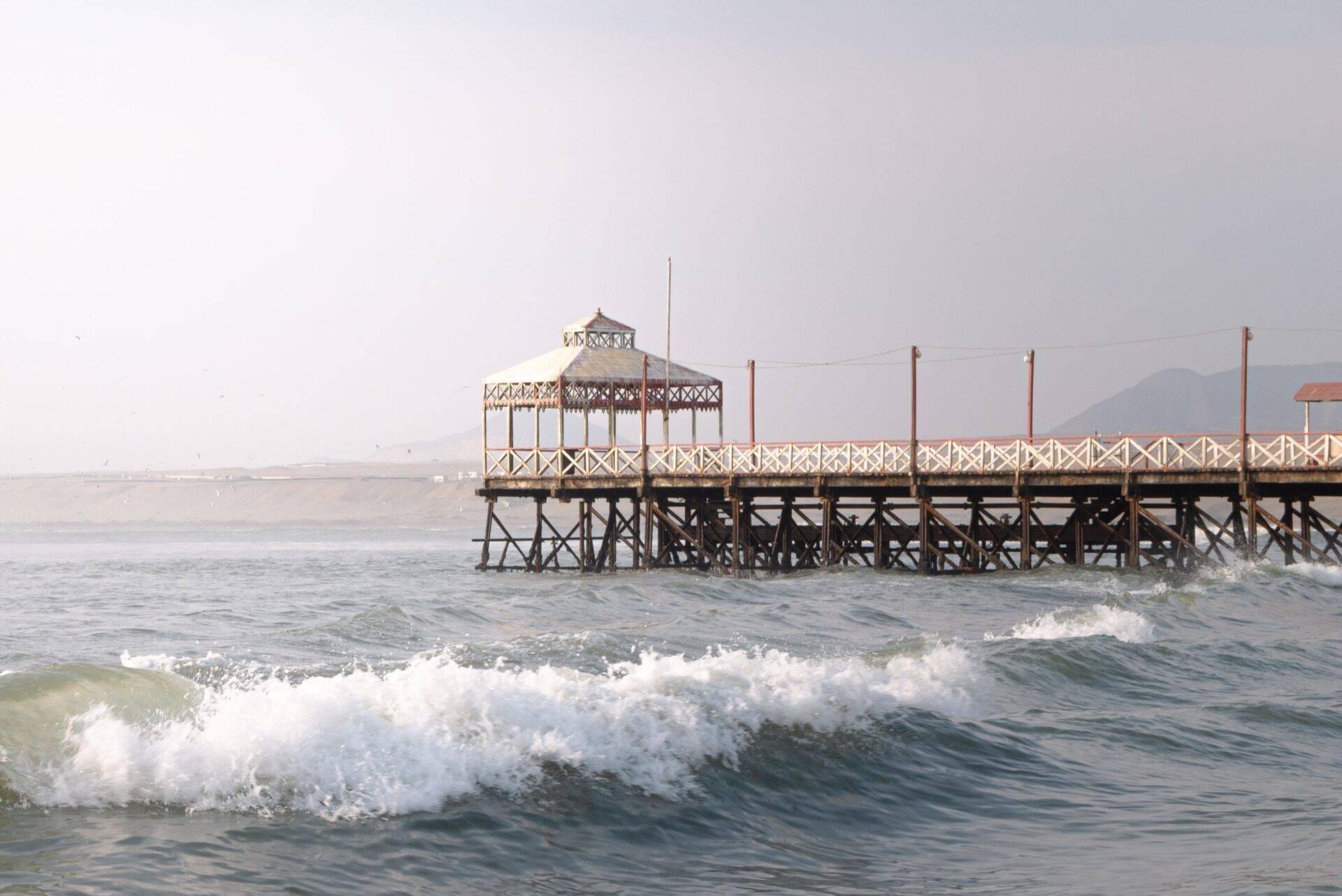 Un muelle de madera con pabellón octogonal frente al océano con olas y montañas al fondo