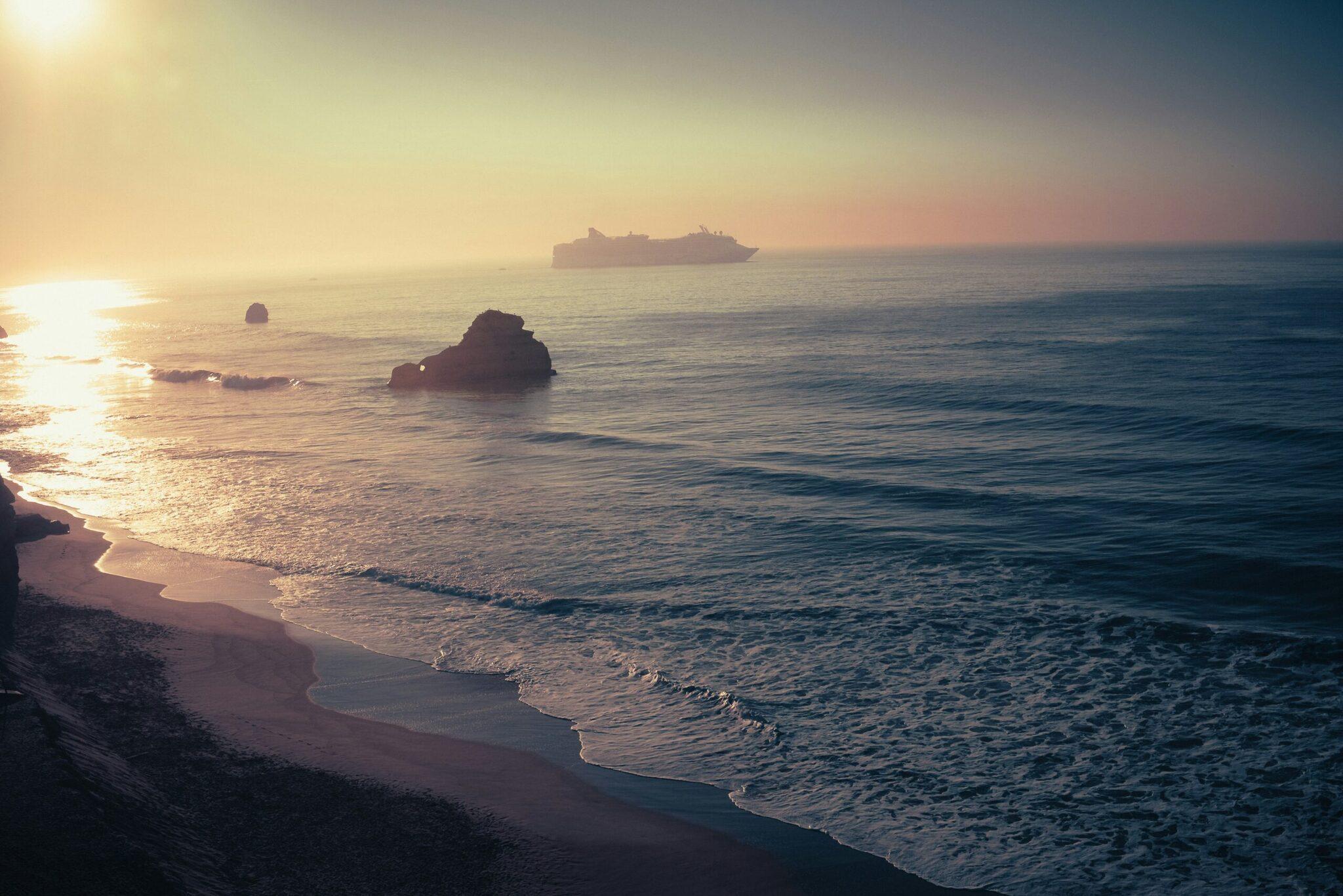 Goldenes Sonnenuntergangslicht über einem felsigen Strand mit einem Kreuzfahrtschiff am Horizont