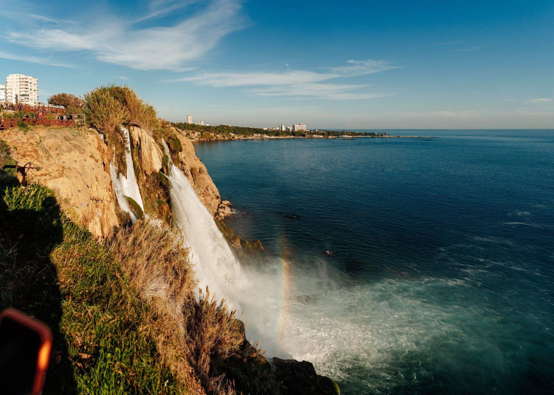 Cascata spettacolare che precipita da una scogliera costiera rocciosa verso il mare turchese mediterraneo sotto un cielo blu sgombro