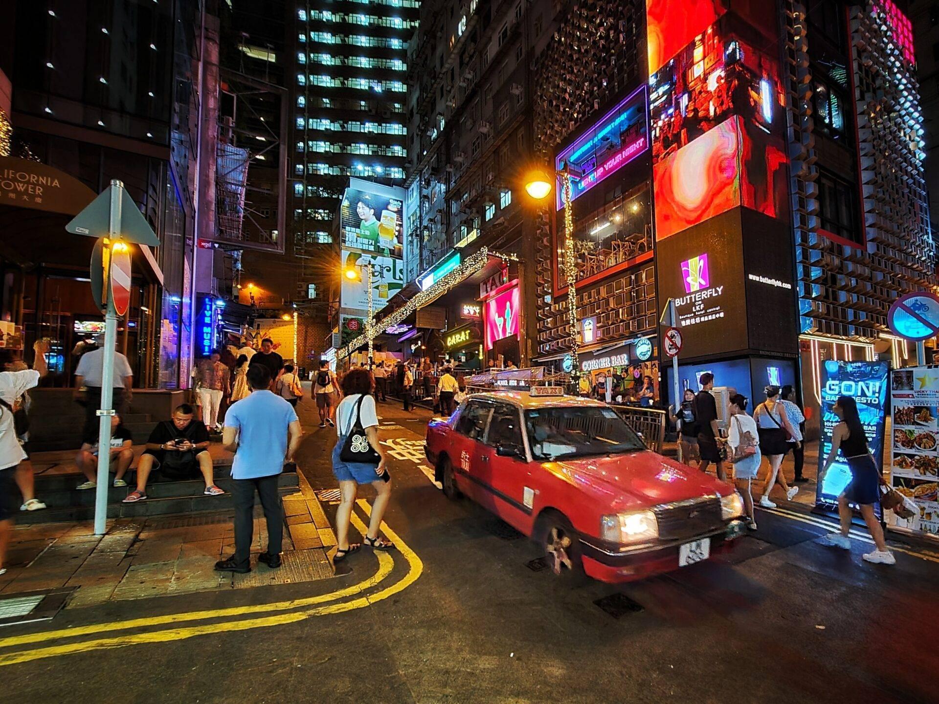 Belebte Fußgängerzone bei Nacht mit beleuchteten Wolkenkratzern, bunten Werbetafeln und einem roten Taxi zwischen Passanten
