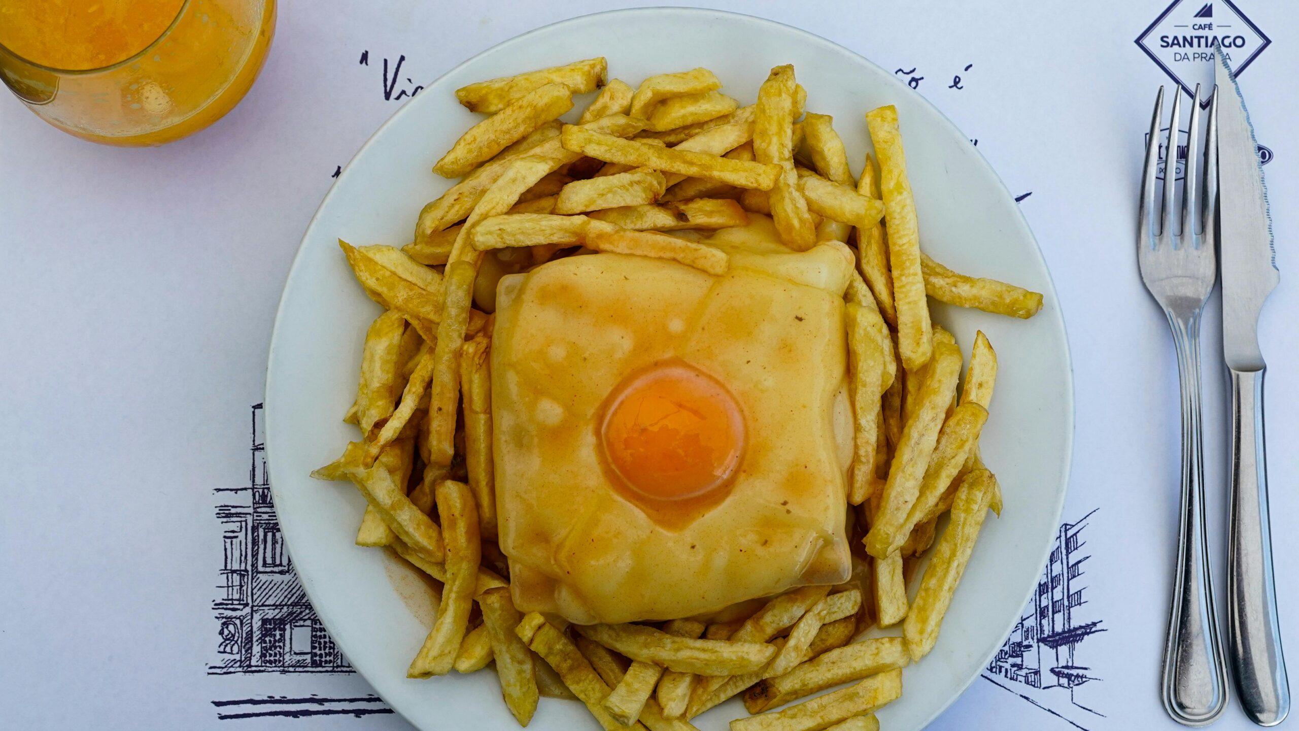 Un sándwich tostado con queso y huevo frito, acompañado de patatas fritas doradas, servido en un restaurante portugués