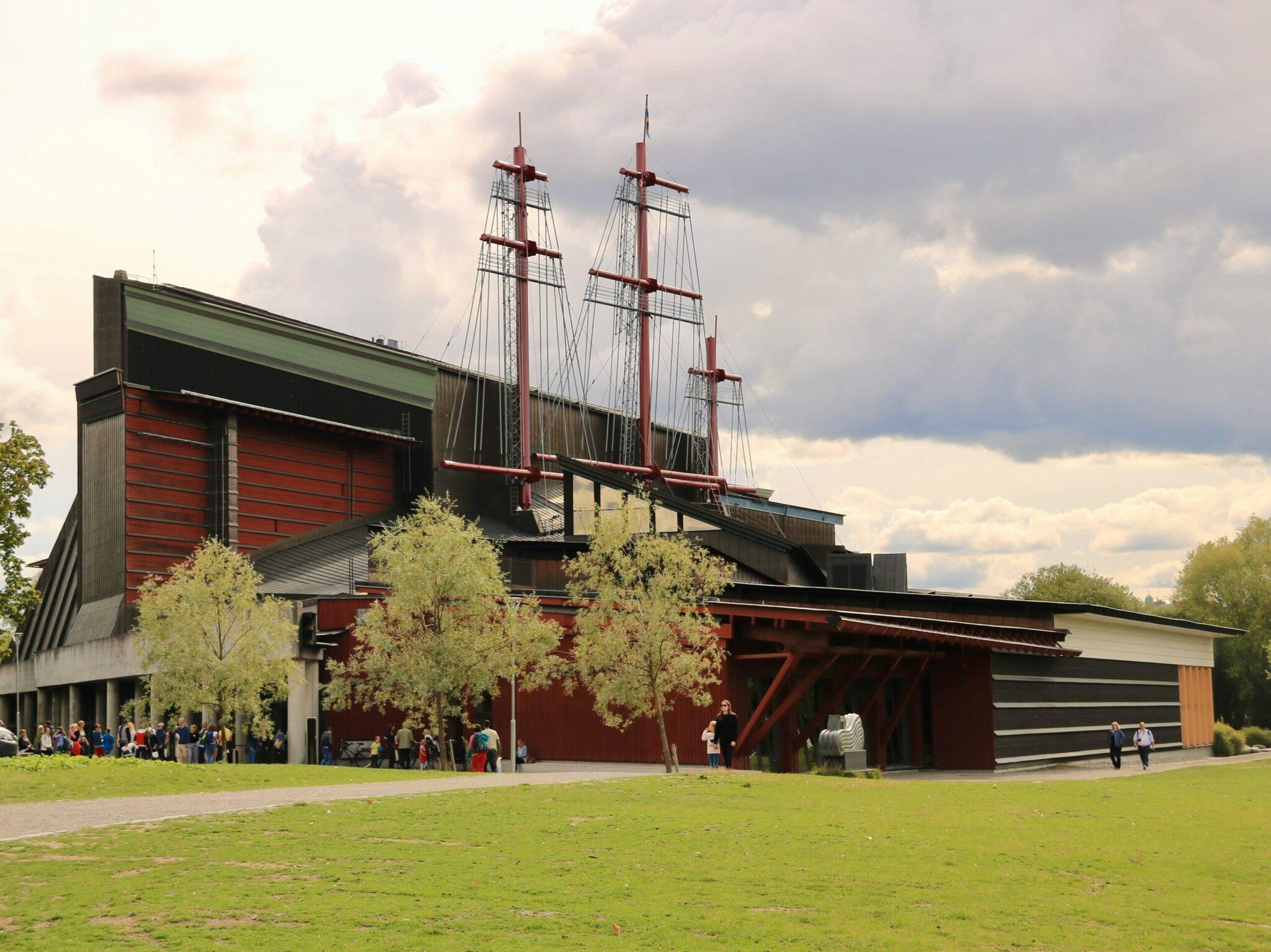 Un museo moderno de fachadas rojas con una réplica de barco de vela integrada en el techo, rodeado de césped y árboles