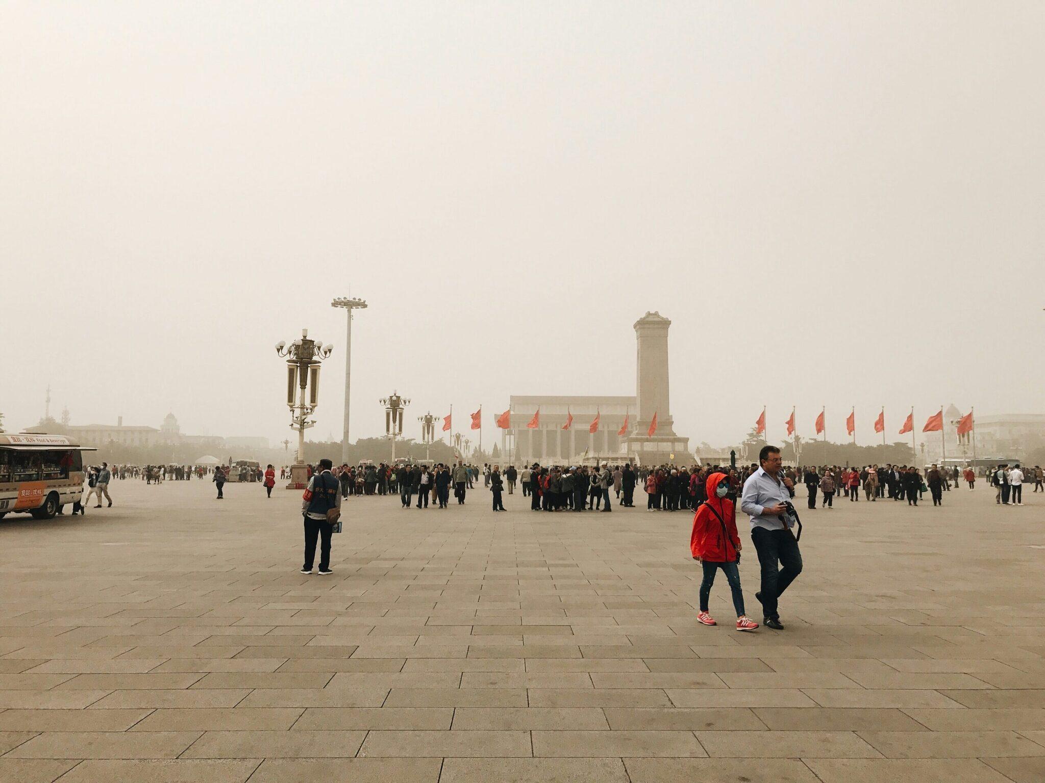 Enorme plaza pavimentada con banderas rojas, un monumento conmemorativo y visitantes en un día neblinoso