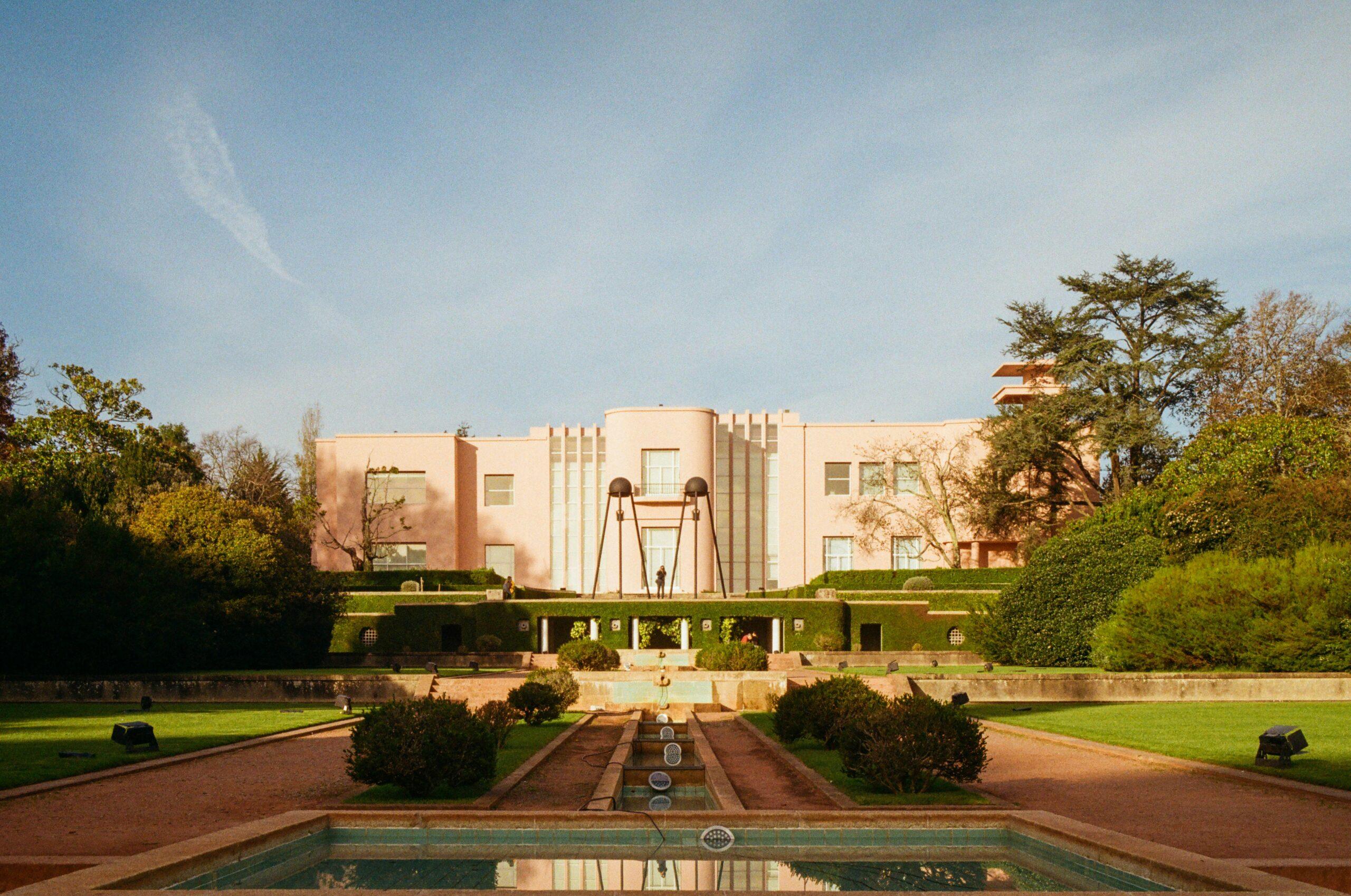 Un edificio Art Decó de color rosa pálido con fuentes y estanque de reflejos, rodeado de jardines verdes y árboles