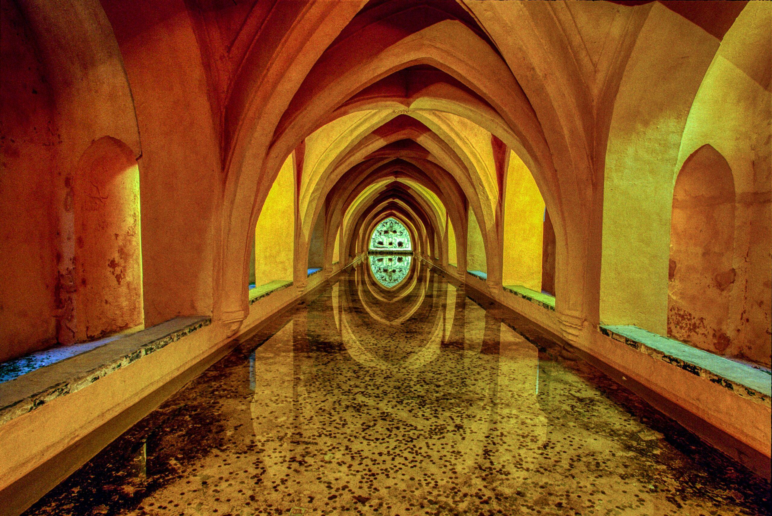 Galería subterránea con bóvedas góticas iluminadas en amarillo y rojo, con el suelo anegado que refleja la arquitectura