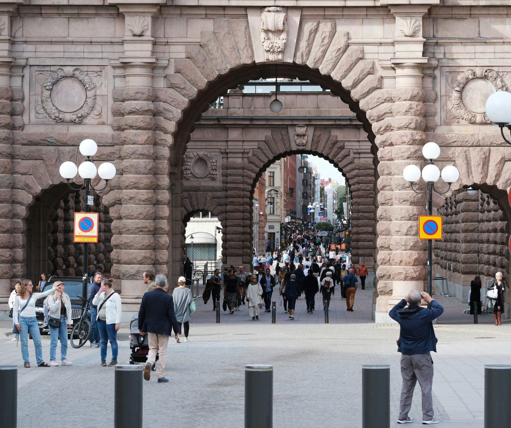 Una calle peatonal animada que atraviesa un imponente arco de piedra histórico con edificios de ladrillo al fondo