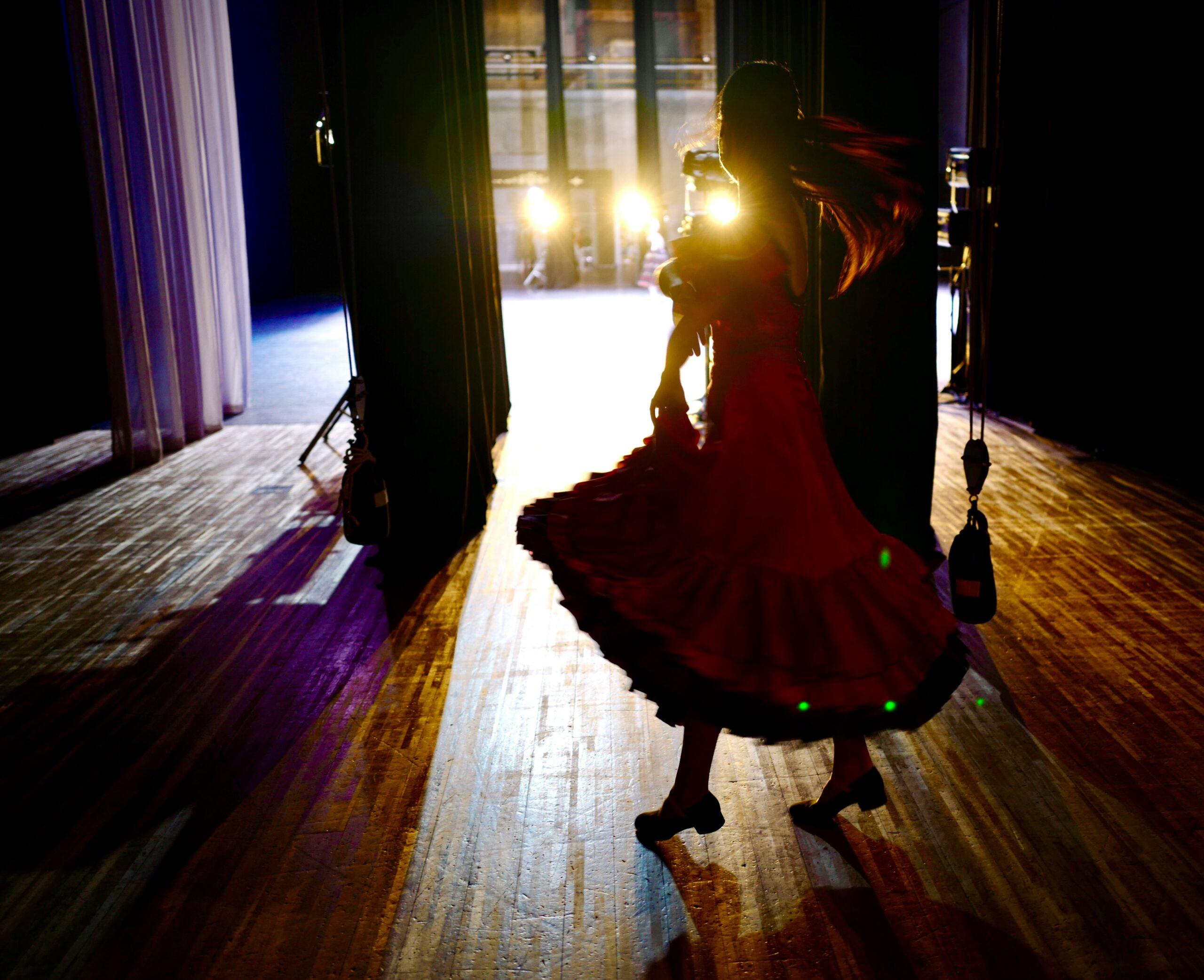 Una mujer con vestido rojo baila en el escenario, iluminada por focos en una sala de espectáculos