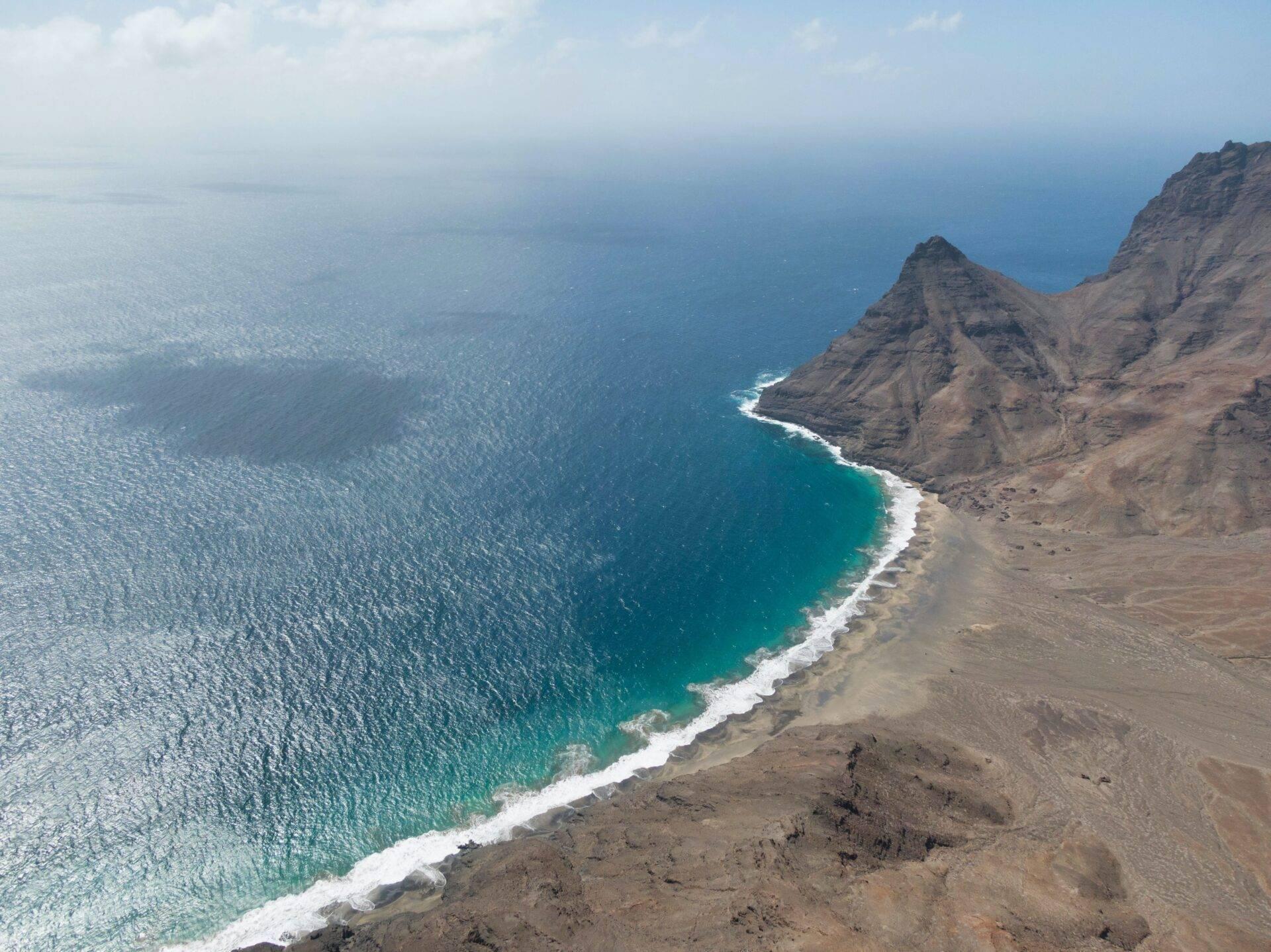 Vue aérienne d’une côte volcanique aride avec falaises brunes, plage de sable noir et océan turquoise aux vagues blanches