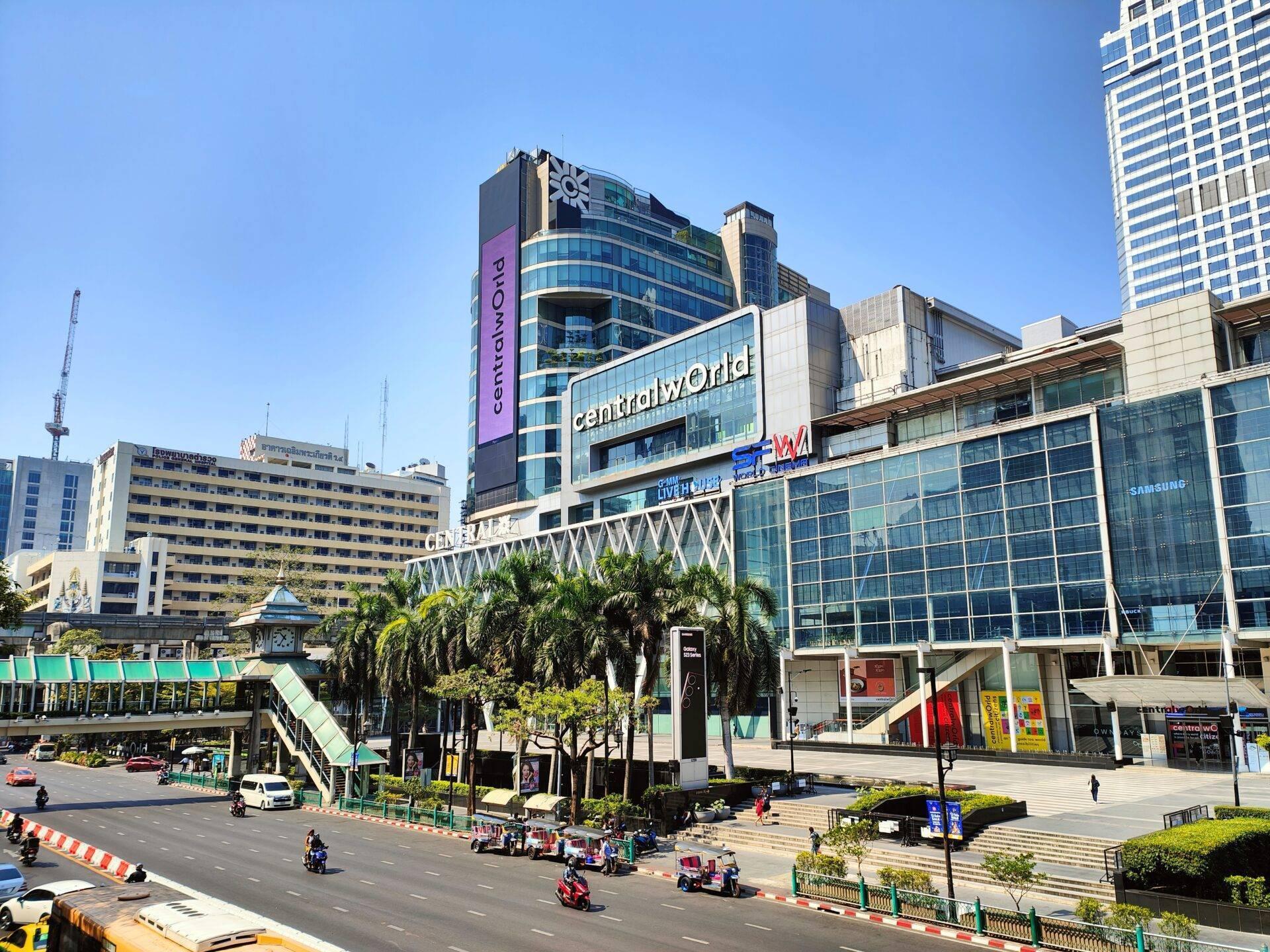 Un’ampia piazza commerciale urbana con moderno centro commerciale, grattacieli, palme e tuk-tuk rossi sotto un cielo azzurro