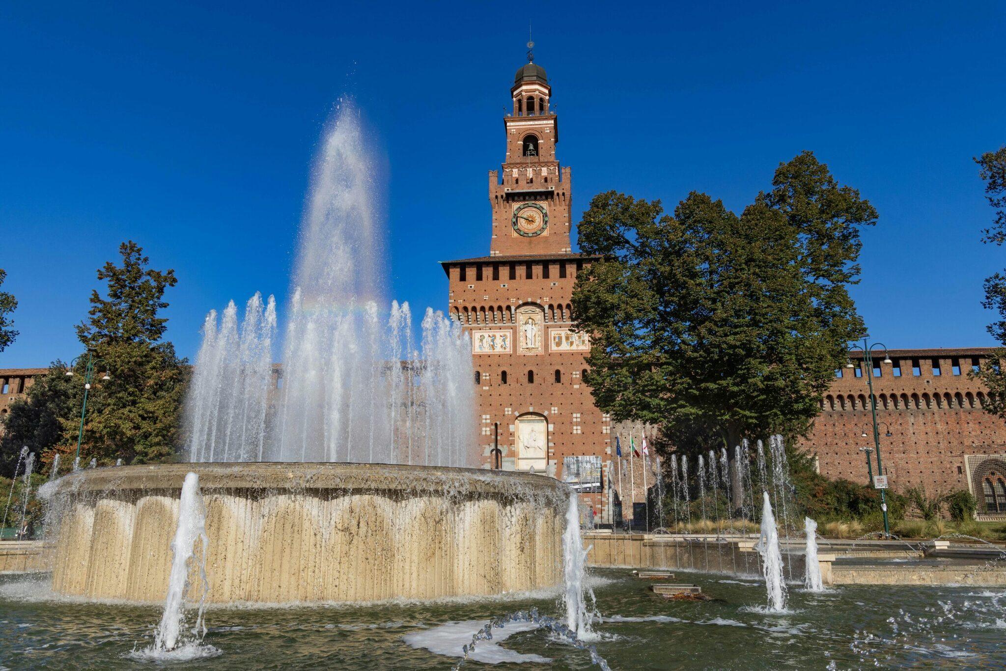 Brunnen und mittelalterliches rotes Backsteinschloss mit Uhrturm unter wolkenlosem blauem Himmel