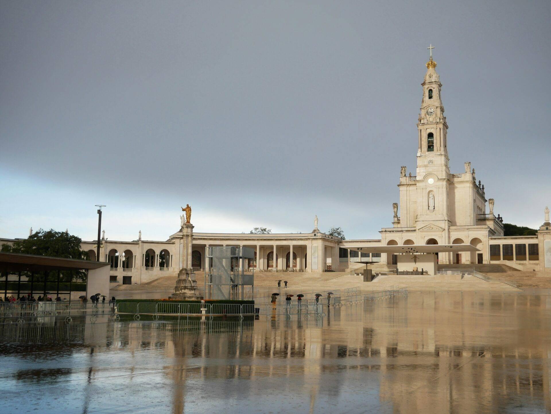 Ein weißes Religionsheiligtum mit Campanile, das eine gepflasterte Esplanade überragt, gesäumt von ruhigem, spiegelndem Wasser