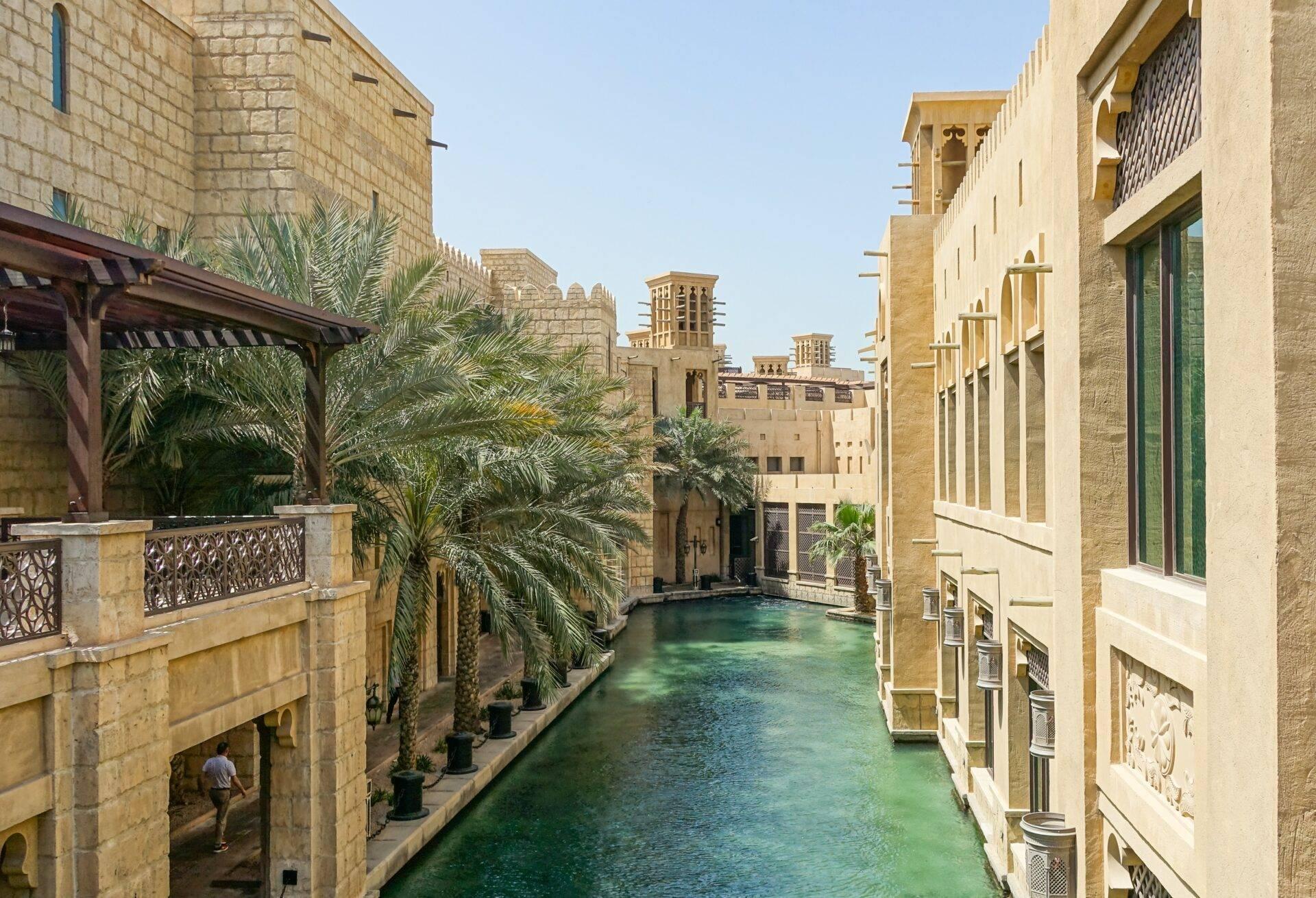 Ein türkisfarbener Wasserkanal gesäumt von ockerfarbenen Steinhäusern mit Palmen und traditioneller Nahost-Architektur