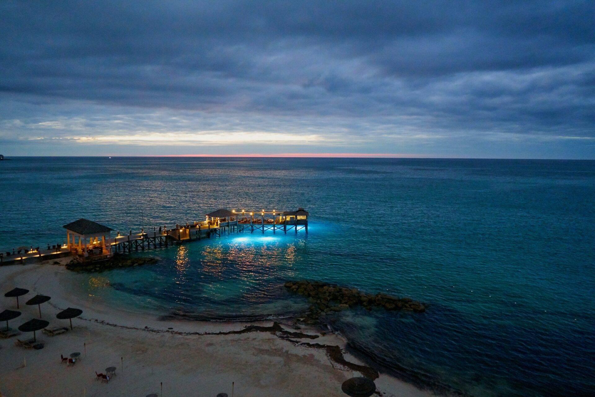 An illuminated pier stretching out into a turquoise sea at dusk, with beach and parasols