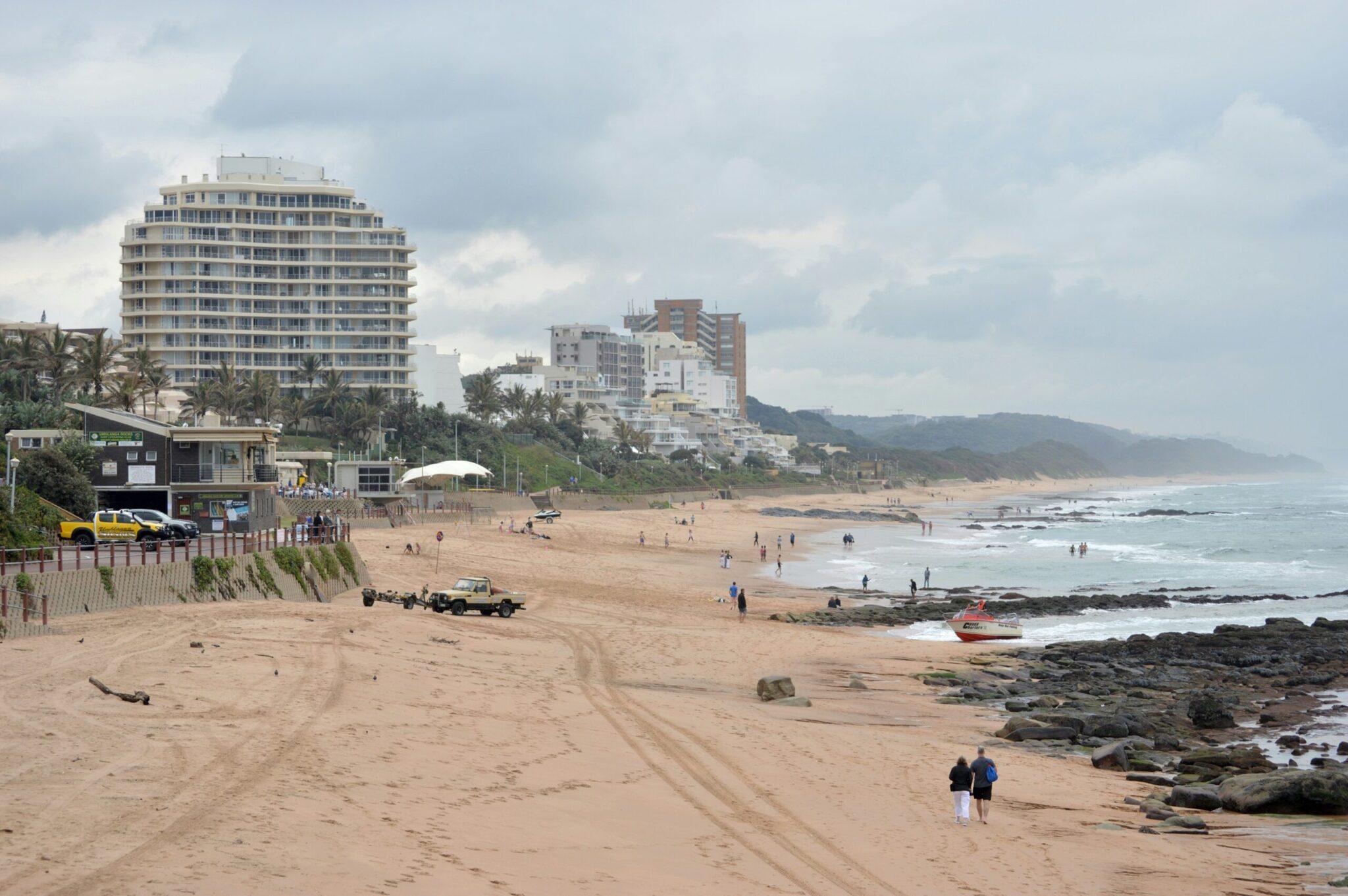 Una playa costera con edificios residenciales, rocas negras, bañistas y vehículos sobre la arena