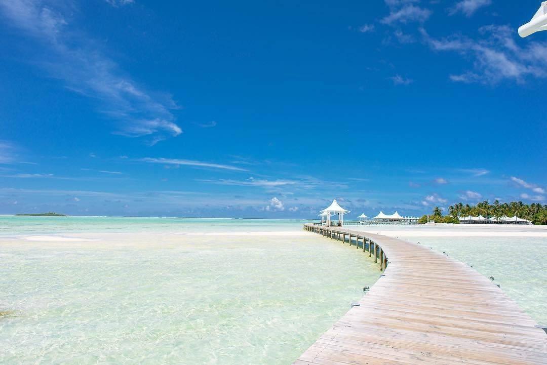 Un pontile di legno si allunga verso un gazebo bianco su una spiaggia di sabbia fine con acque turchesi cristalline