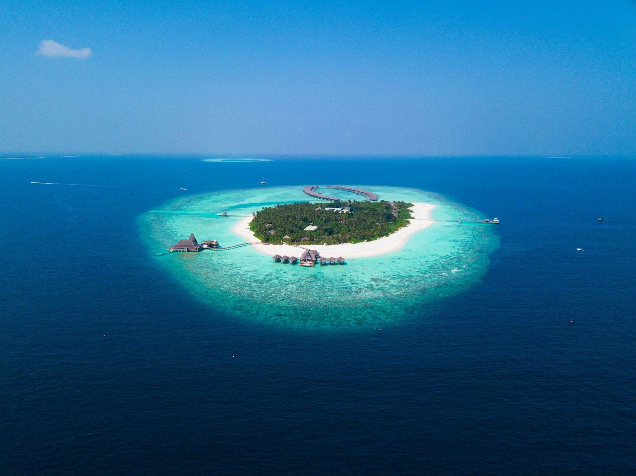Luftaufnahme einer kreisrunden Tropeninsel mit weißem Sandstrand, türkisem Lagunenring und Überwasserbungalows