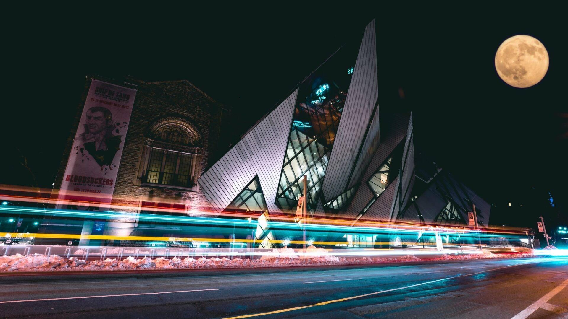 Nächtliche Außenansicht eines modernen Museums mit beleuchteter Winkelarchitektur, Vollmond und Lichtspuren von Fahrzeugen