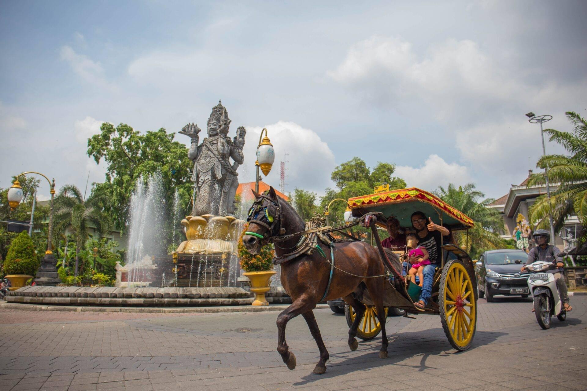Un carro tradizionale giallo e rosso trainato da cavalli passa davanti a una statua divina in una piazza pubblica indonesiana