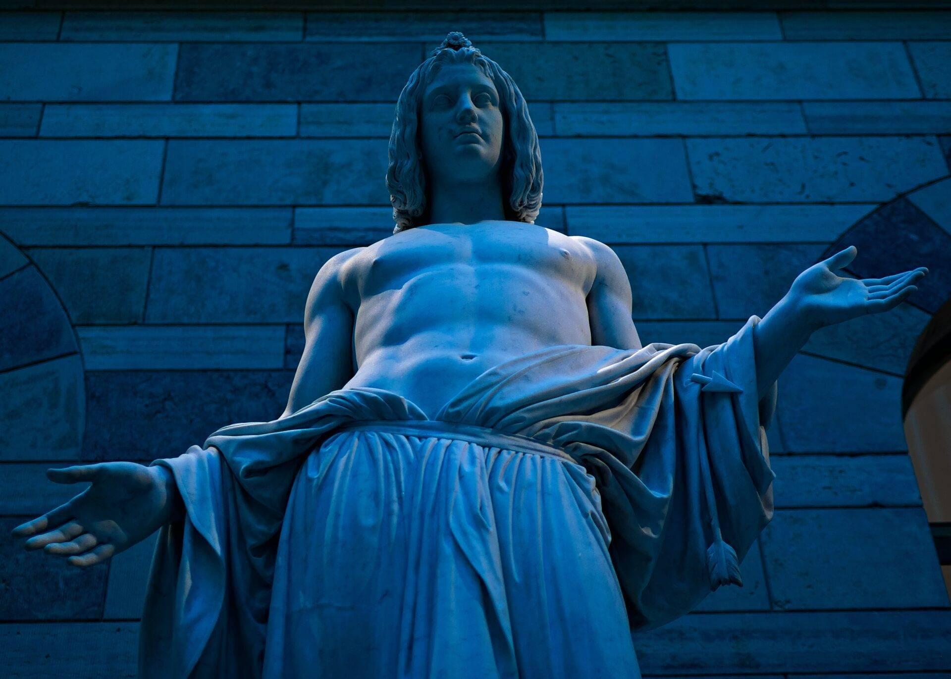 Marmorstatue im blauen Nachtlicht vor einer Steinwand, nachdenkliche Pose mit klassischem Gewand