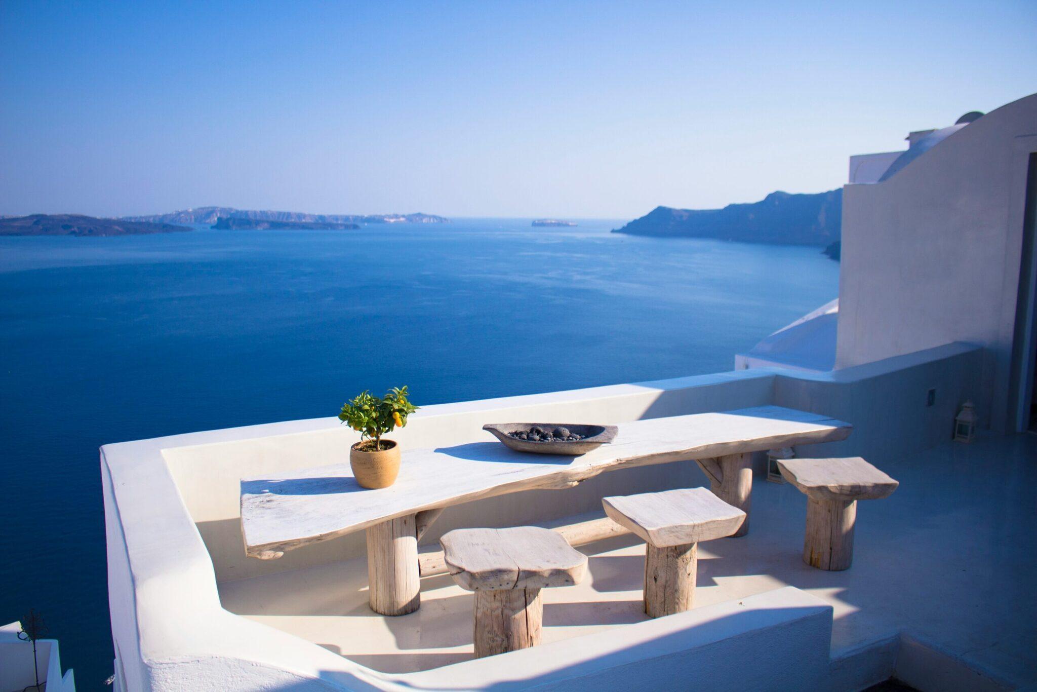 Terrazza bianca affacciata su una baia mediterranea con tavolo in legno, sgabelli e vista sulle isole rocciose