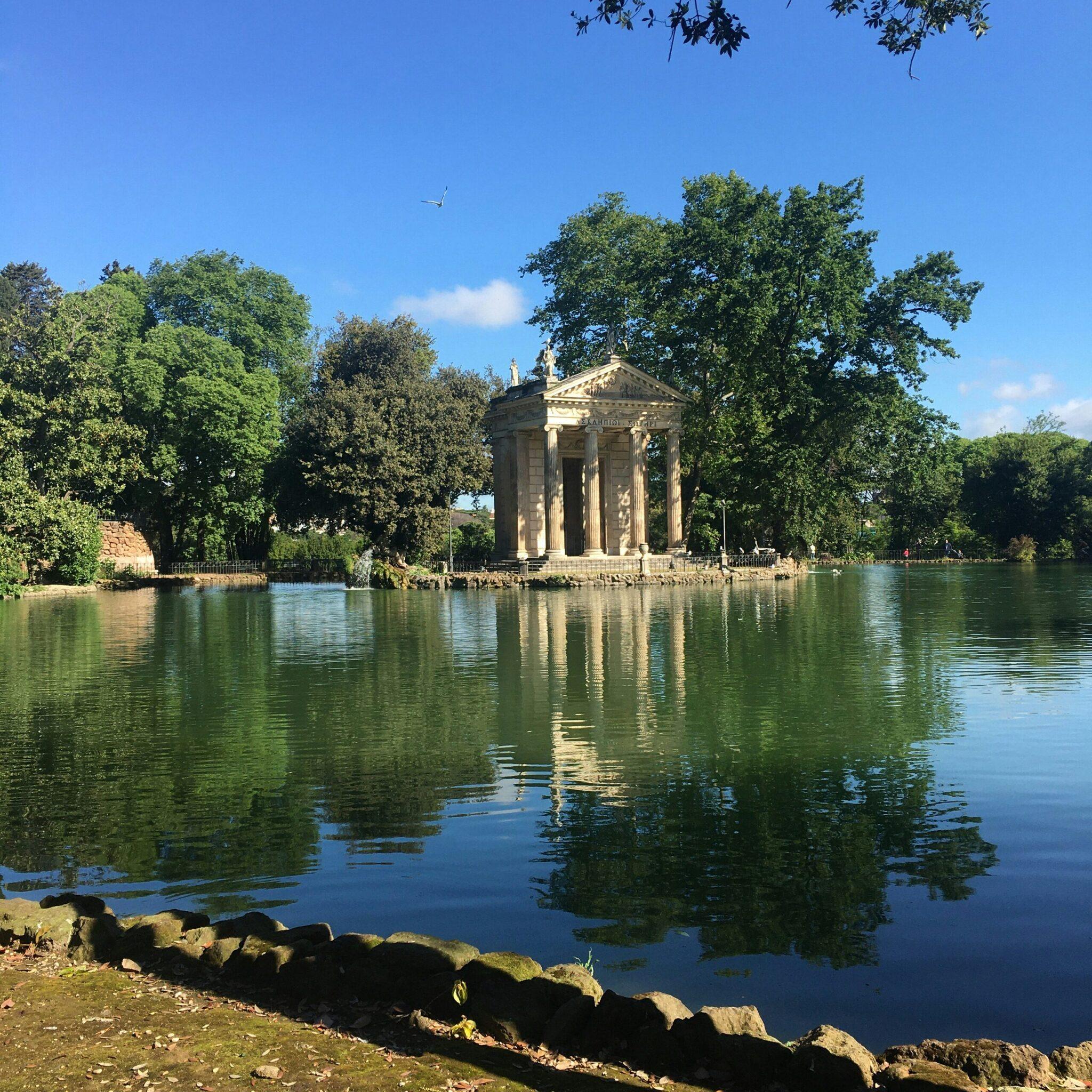 Templo clásico de columnas reflejándose en un lago tranquilo, rodeado de vegetación exuberante en un día soleado