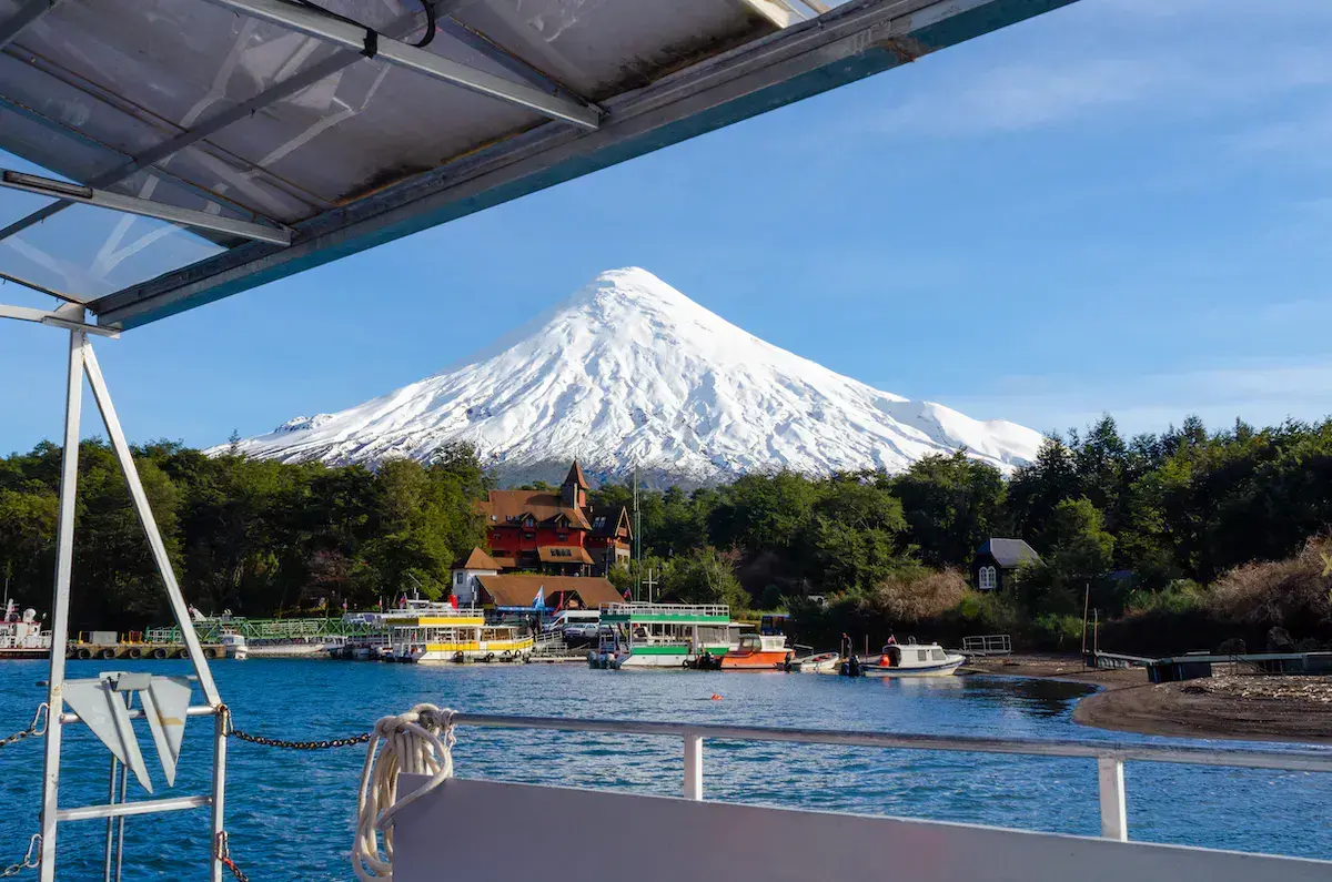 Vista desde un barco de un volcán nevado dominando un lago con edificios de colores y embarcaciones