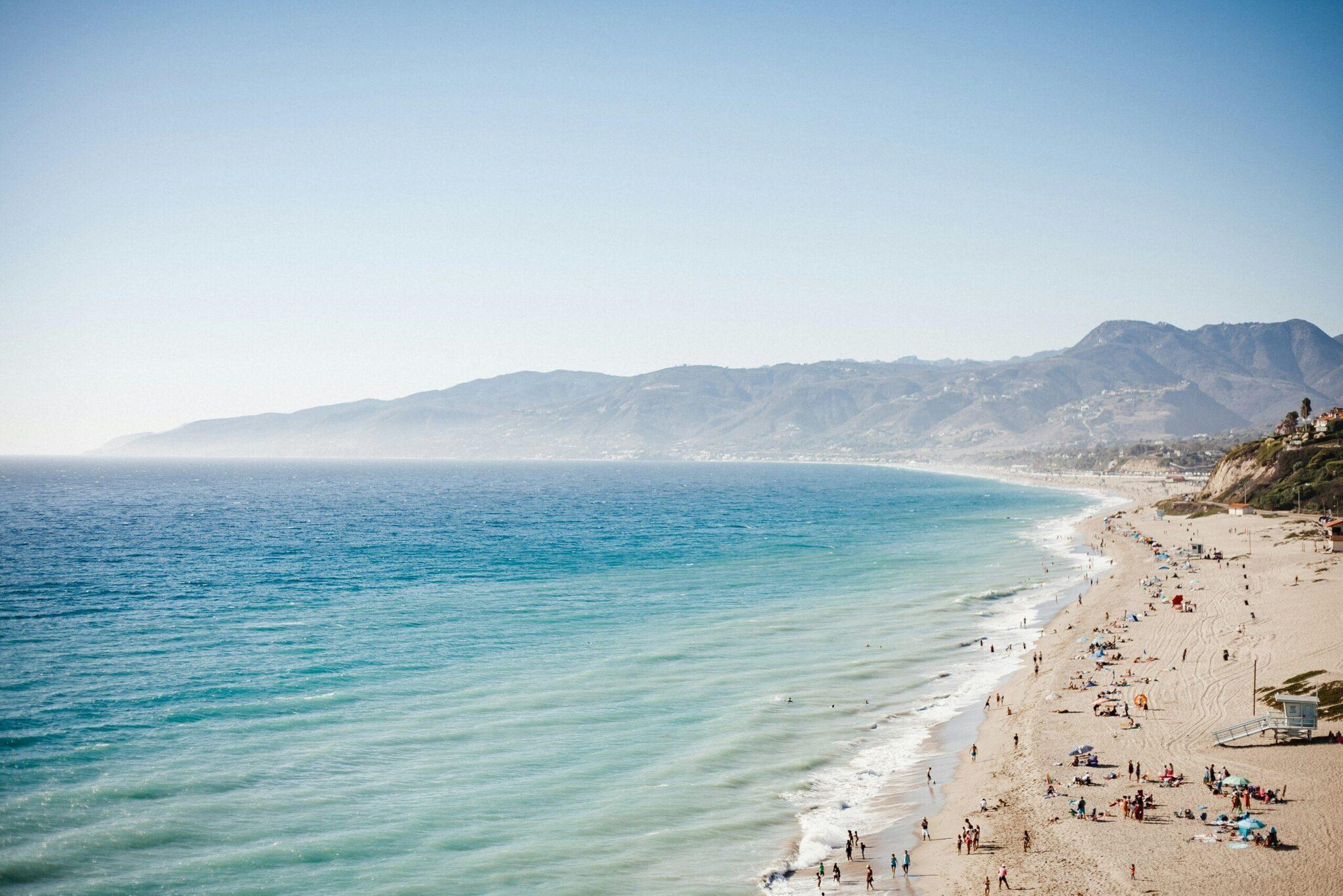 Una larga playa de arena blanca con bañistas frente a un océano turquesa y montañas costeras boscosas