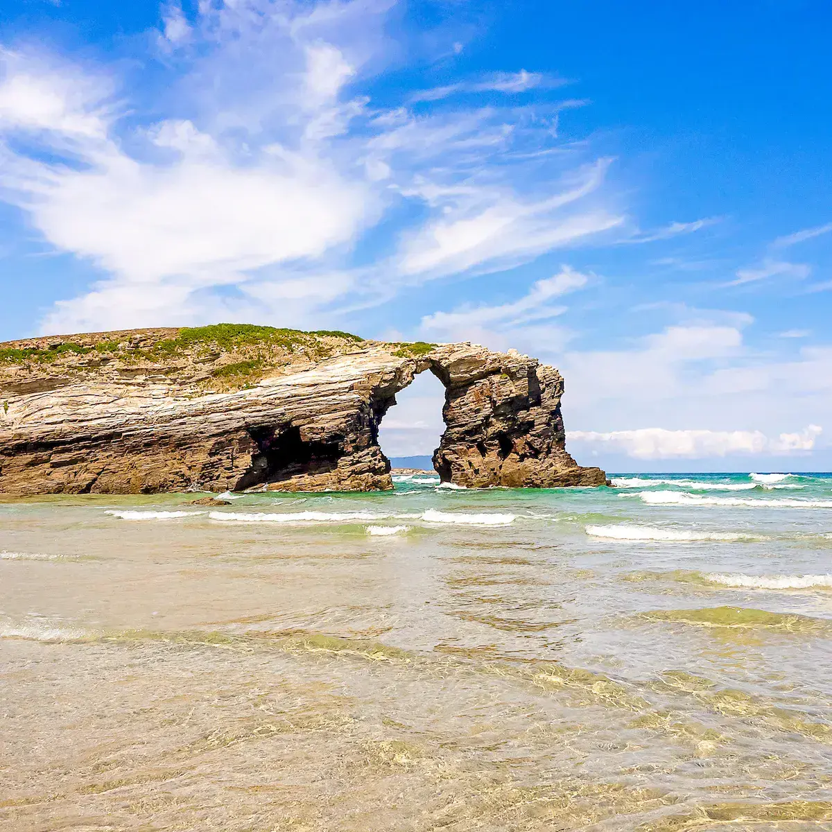 Ein natürlicher Felsbogen ragt aus dem Ozean an einem Feinsandstrand unter wolkenlosem blauem Himmel
