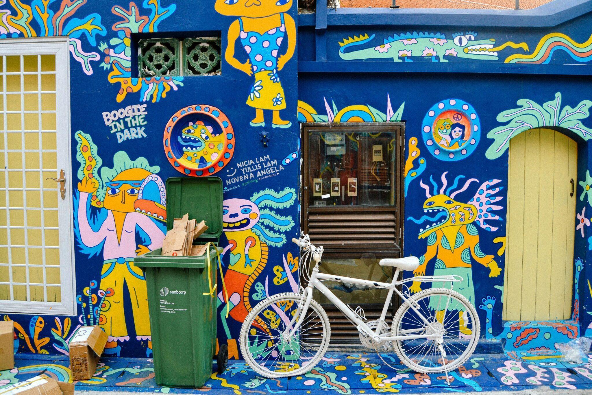 Una bicicletta bianca davanti a una facciata blu ornata da murales colorati e motivi tropicali stilizzati