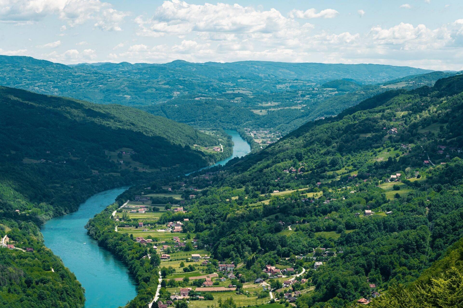 Ein grünes Flusstal, durchzogen von einem türkisfarbenen Fluss, umgeben von bewaldeten Bergen und kleinen Dörfern