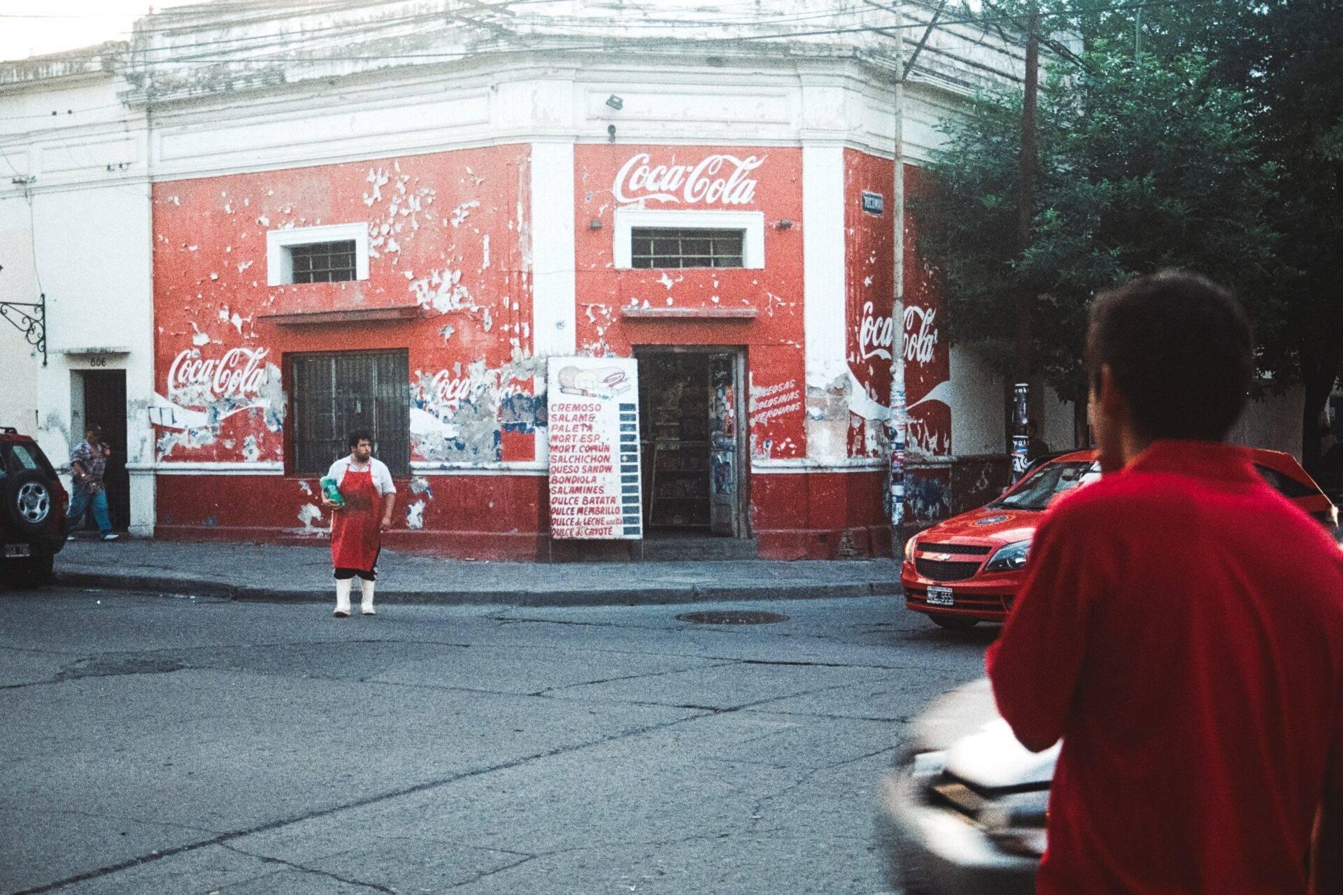 Una strada di un quartiere popolare latinoamericano con un edificio dai muri rossi dipinti con loghi pubblicitari vintage