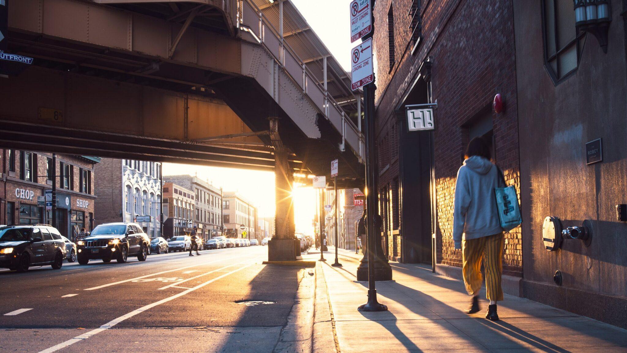 Amerikanische Stadtstraße unter einem Eisenbahnviadukt bei Sonnenuntergang, mit Fußgängern und Autos zwischen Backsteingebäuden