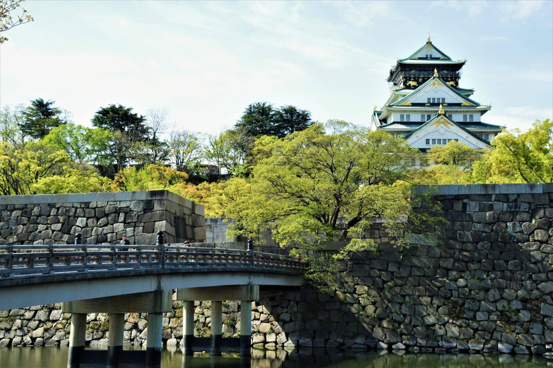 Eine traditionelle japanische Burg mit geschwungenen Dächern überragt eine grüne Landschaft, die von einer Steinbrücke durchquert wird