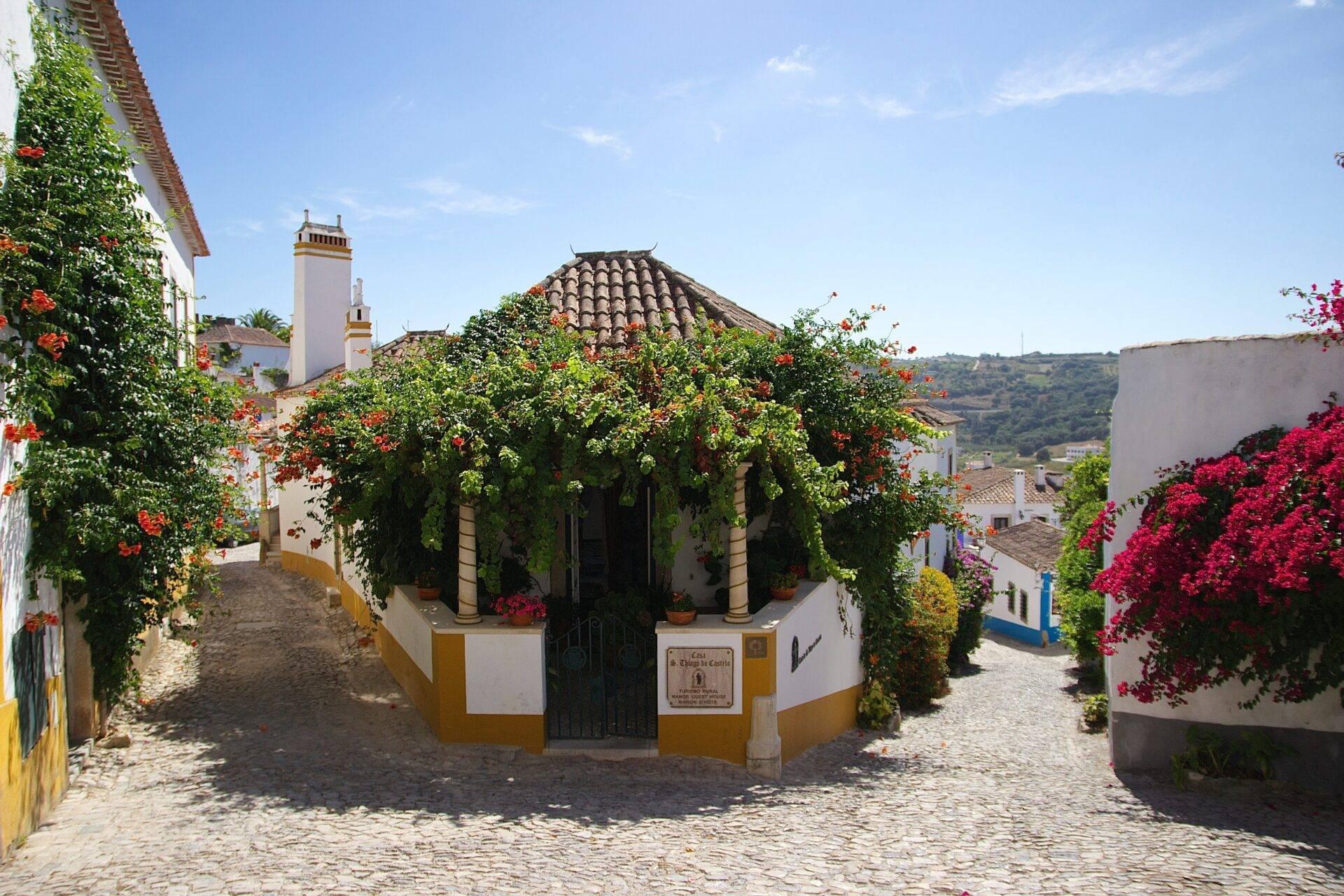 Eine malerische gepflasterte Gasse mit einer weiß-gelben Kapelle, umgeben von leuchtend roten Blüten